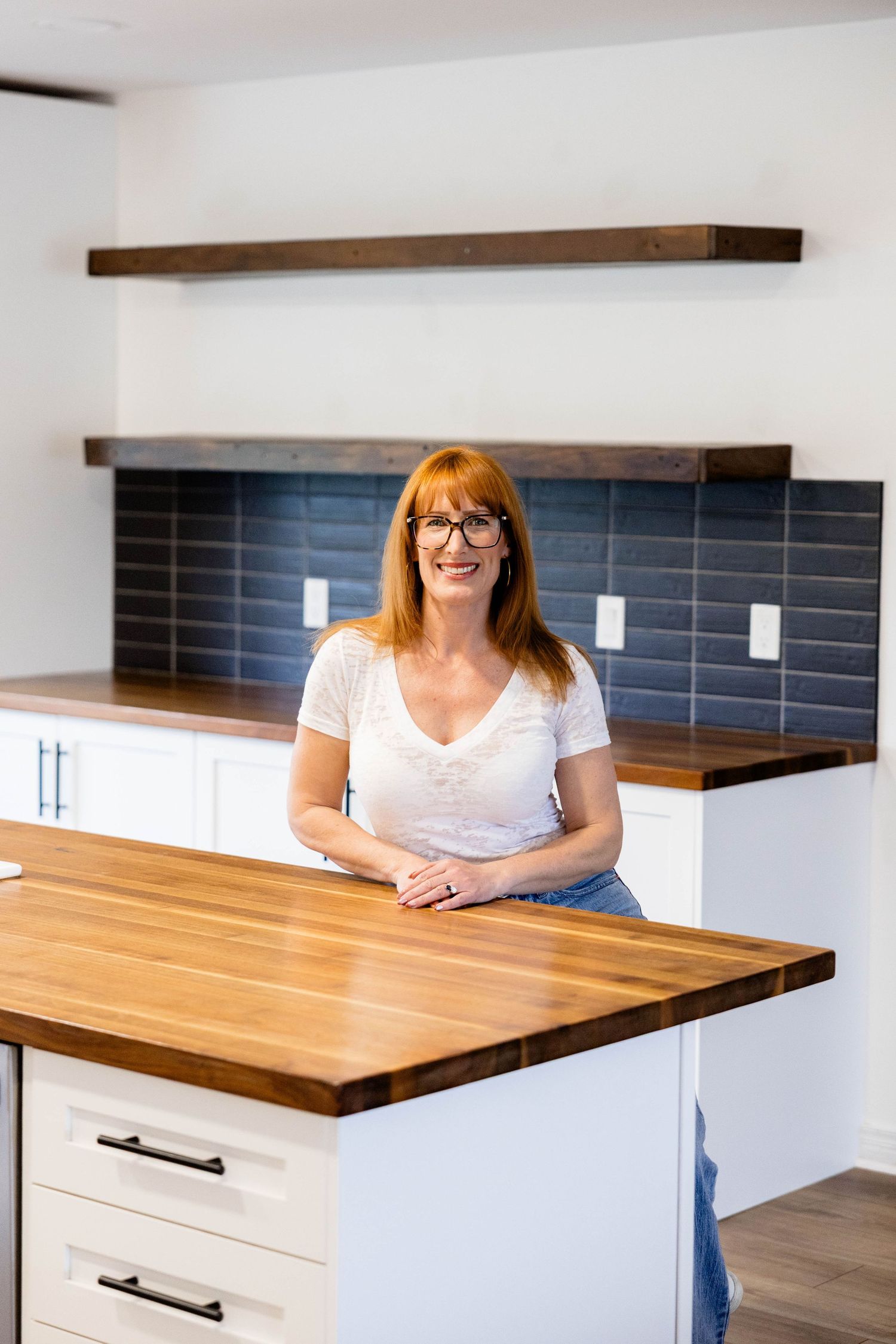 Red-haired woman smiling at kitchen island with wood countertop, white cabinets, and blue subway tile.