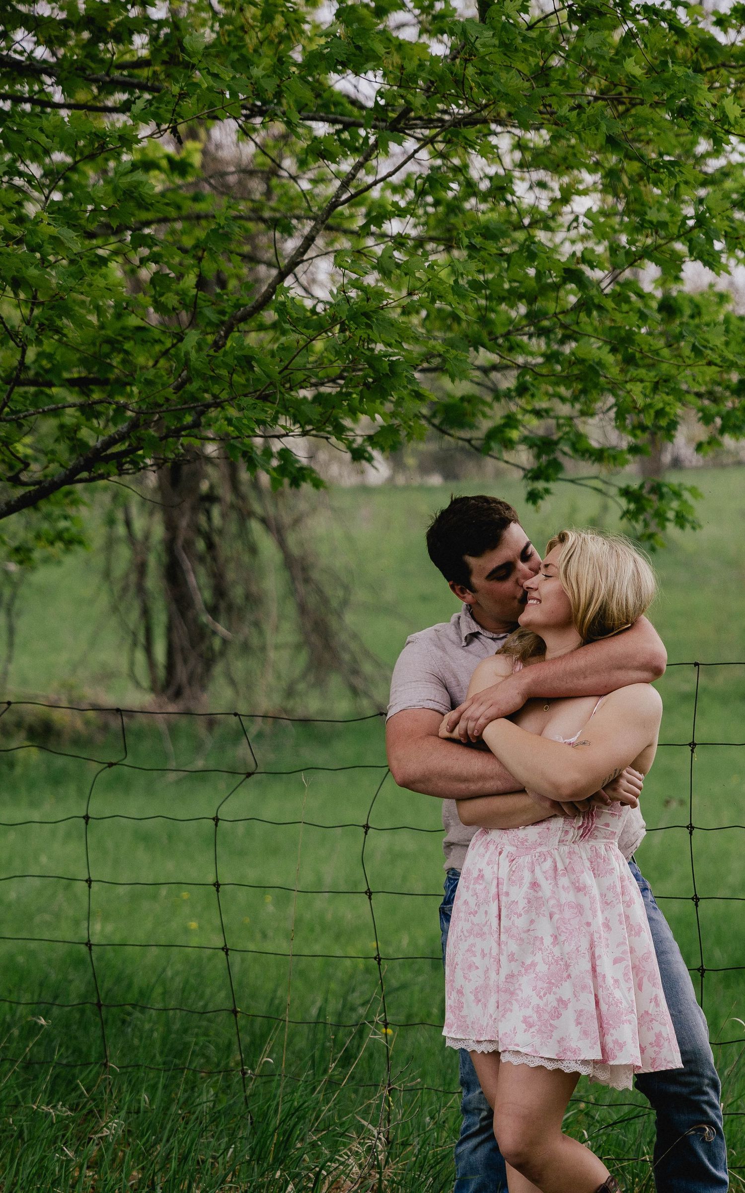 Loving couple embracing and smiling by a wire fence surrounded by lush green trees and grass.