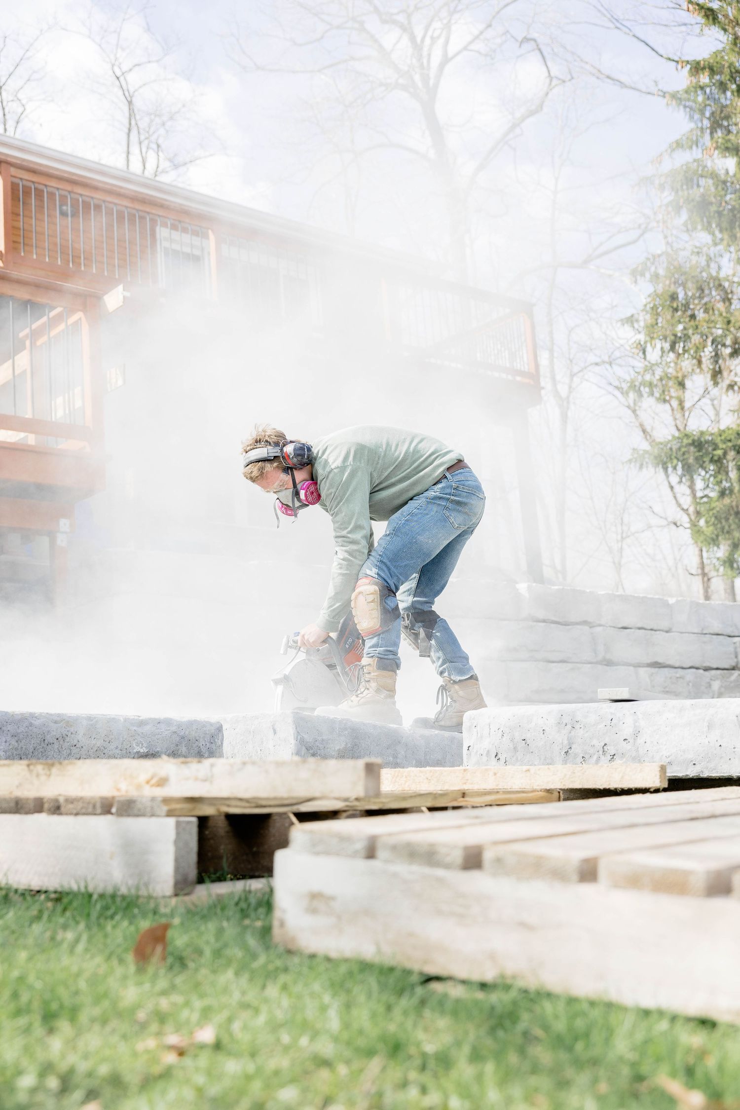 Worker using an angle grinder on concrete blocks with dust cloud near a house.