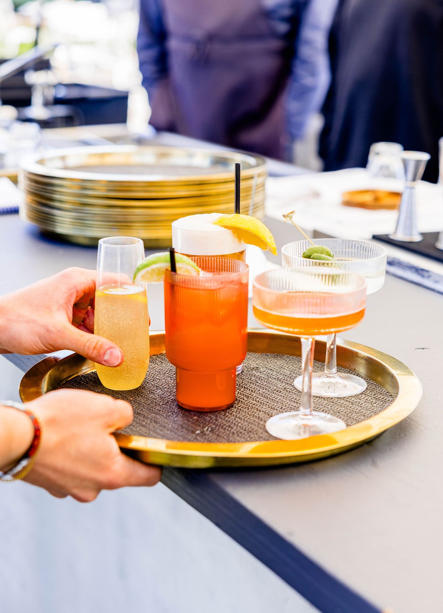 Server holding gold tray with assorted colorful cocktails including a Bloody Mary at outdoor bar.