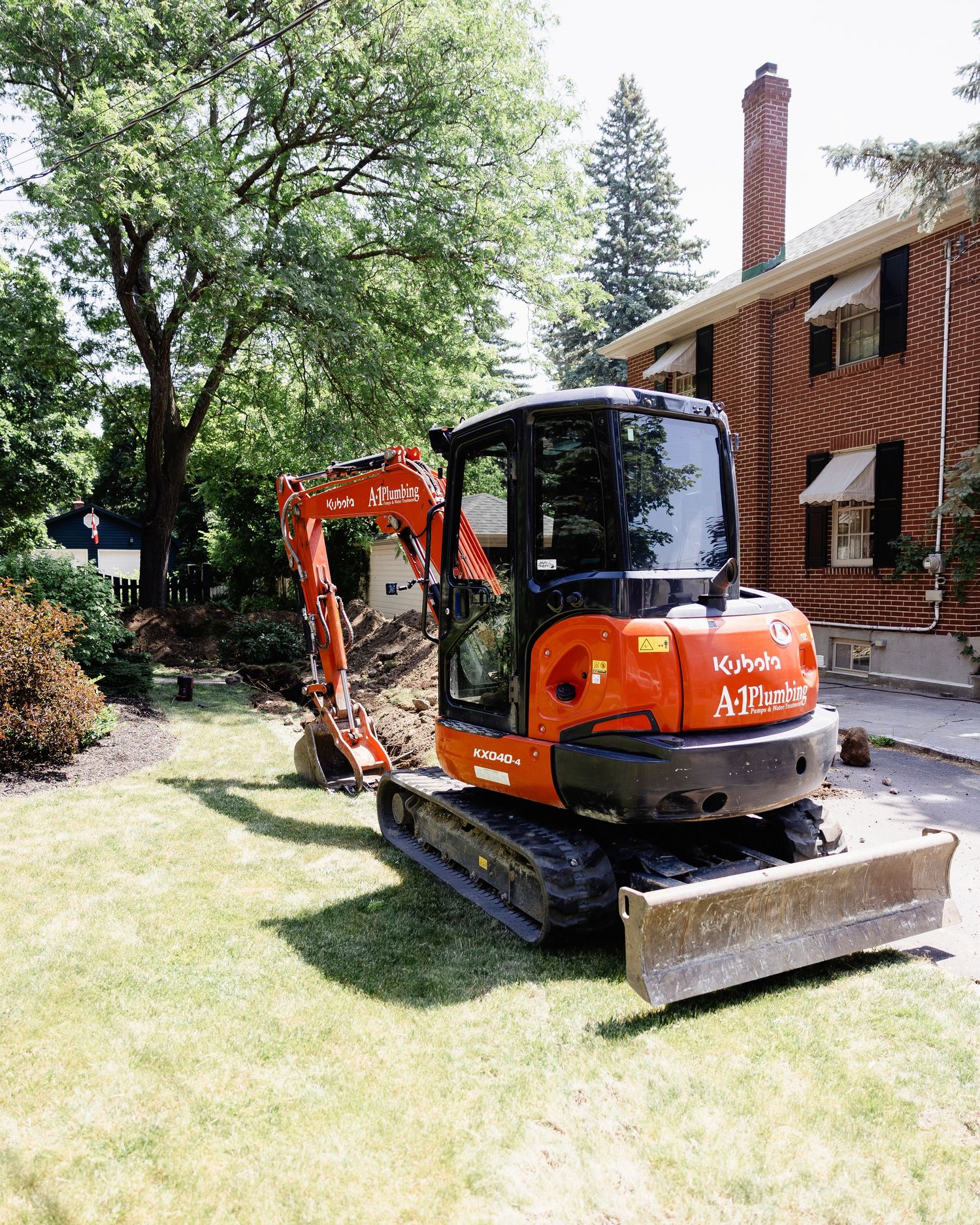 Orange Kubota compact excavator parked on grass beside brick house during landscaping project.