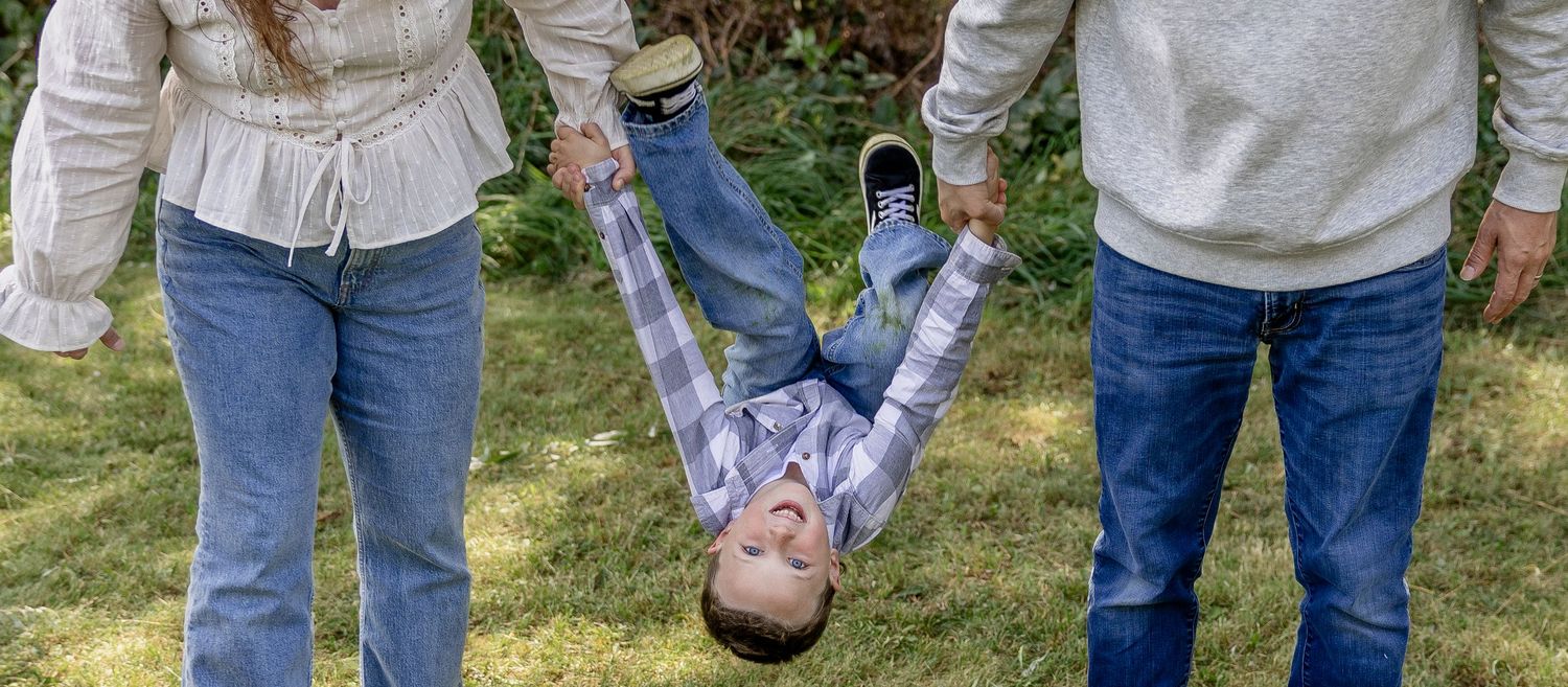 Two parents joyfully swing their young child between them outdoors on a green lawn during a family photo session.