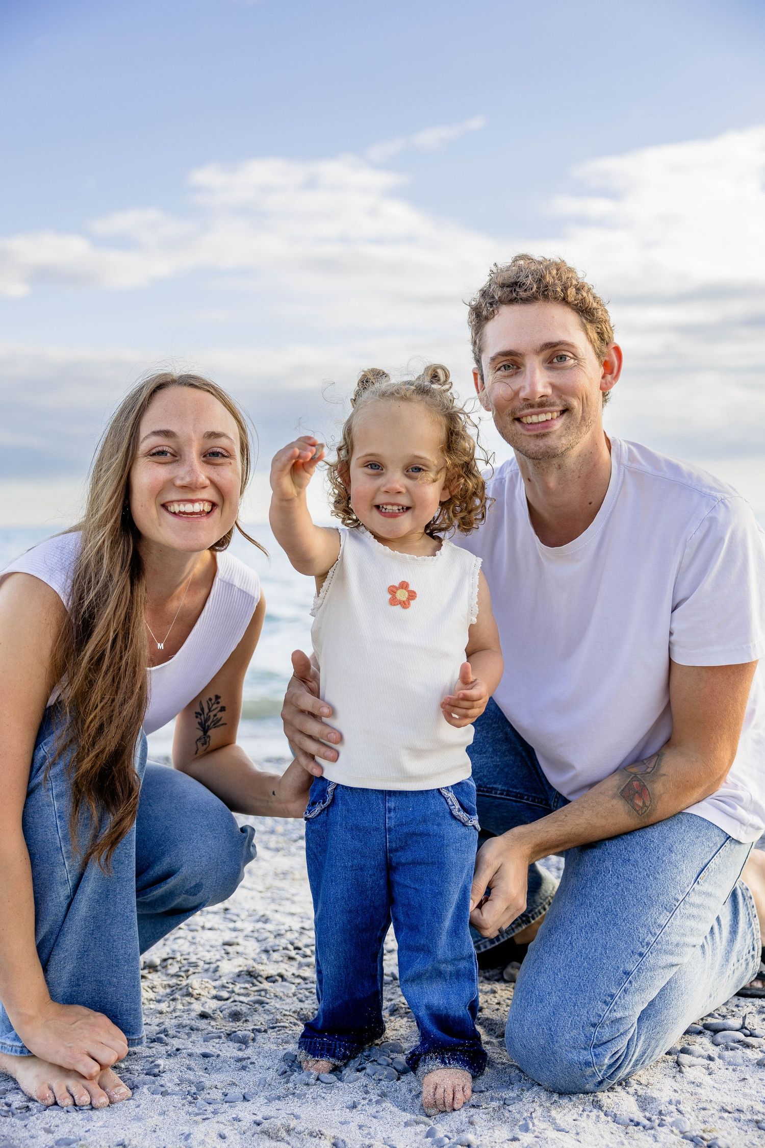 A happy young family of three poses on a sandy beach, with parents in white tees and jeans flanking their smiling toddler.