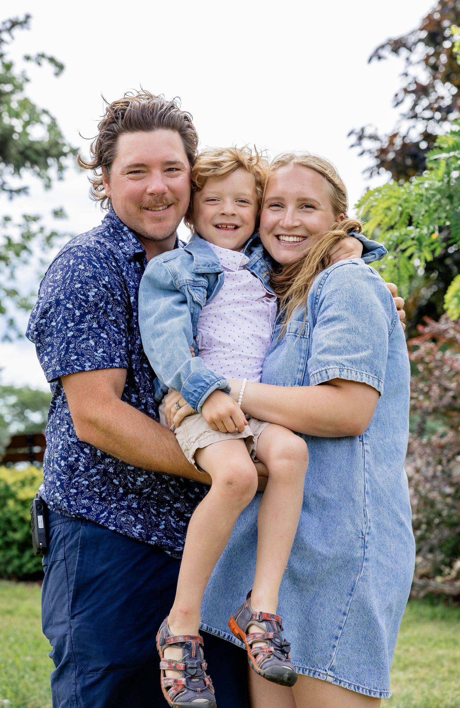 Happy family of three posing outdoors, parents holding young child in garden setting on a sunny day.