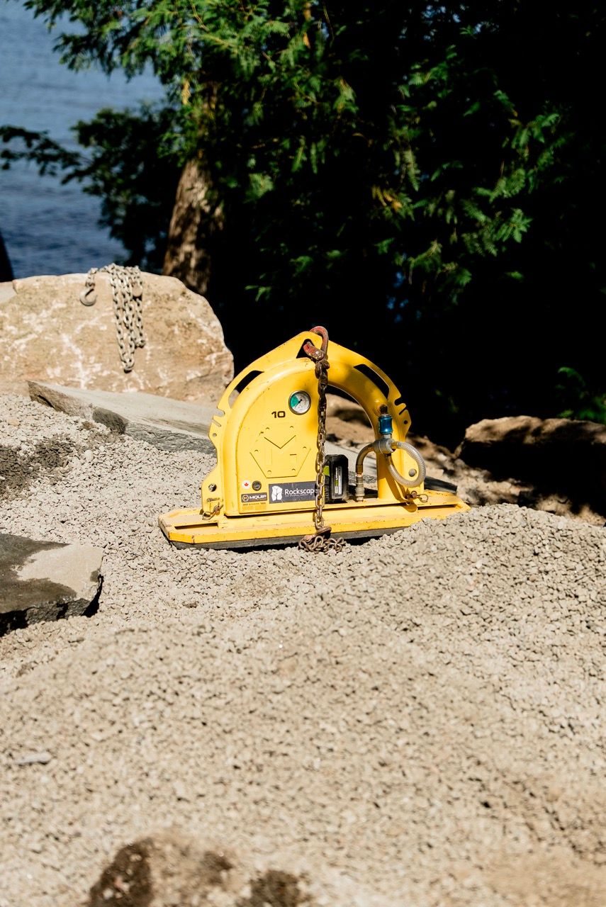 Yellow MQUIP stone lifting clamp attachment sitting on granite rock surface at outdoor construction site.