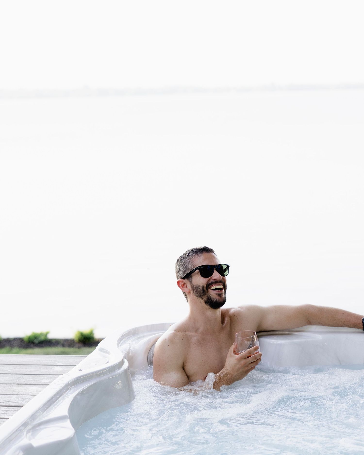 A smiling man wearing sunglasses relaxes in an outdoor hot tub holding a drink with a lake view.