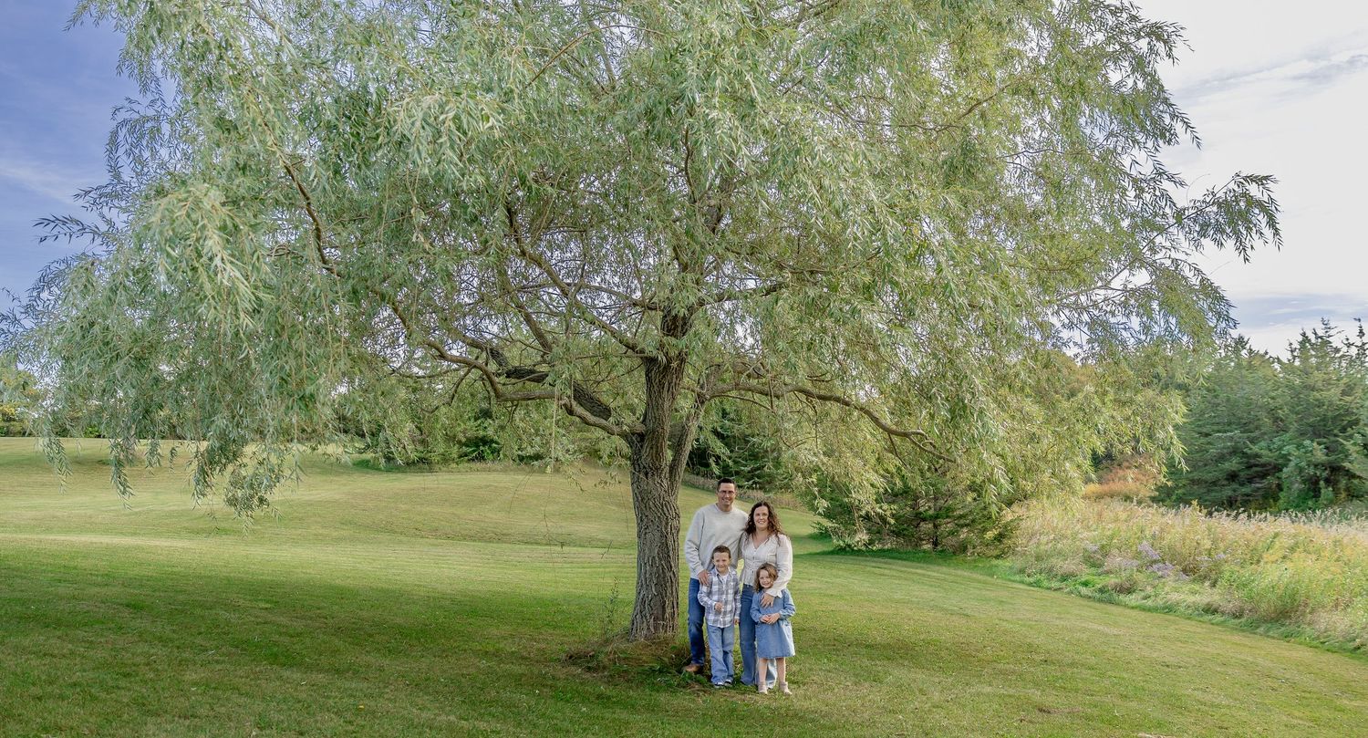 Family posing under a large willow tree on a lush green lawn in a serene outdoor setting.