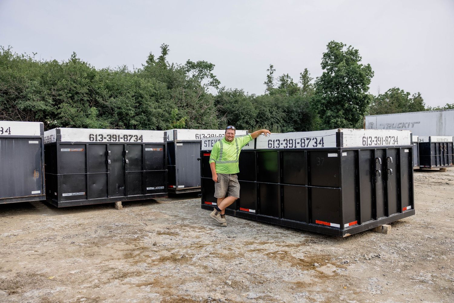 Man in green vest standing beside large black dumpster containers in a gravel yard with phone number 613-781-8234.