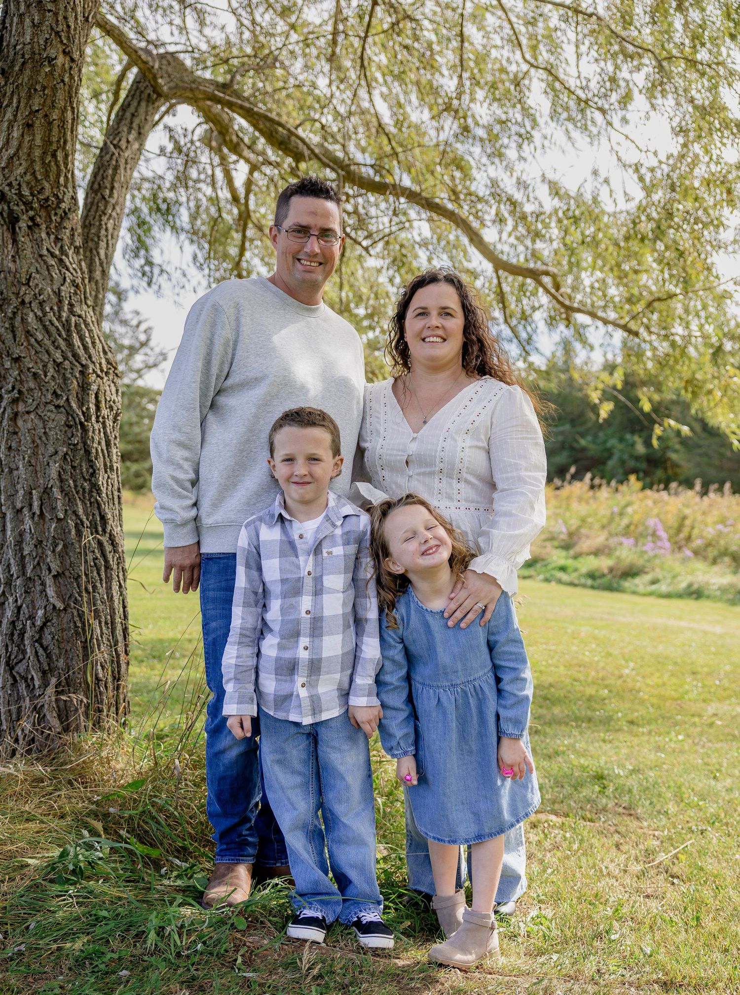 Family of four posing outdoors under a tree, parents with two young children smiling in coordinated blue and white outfits.