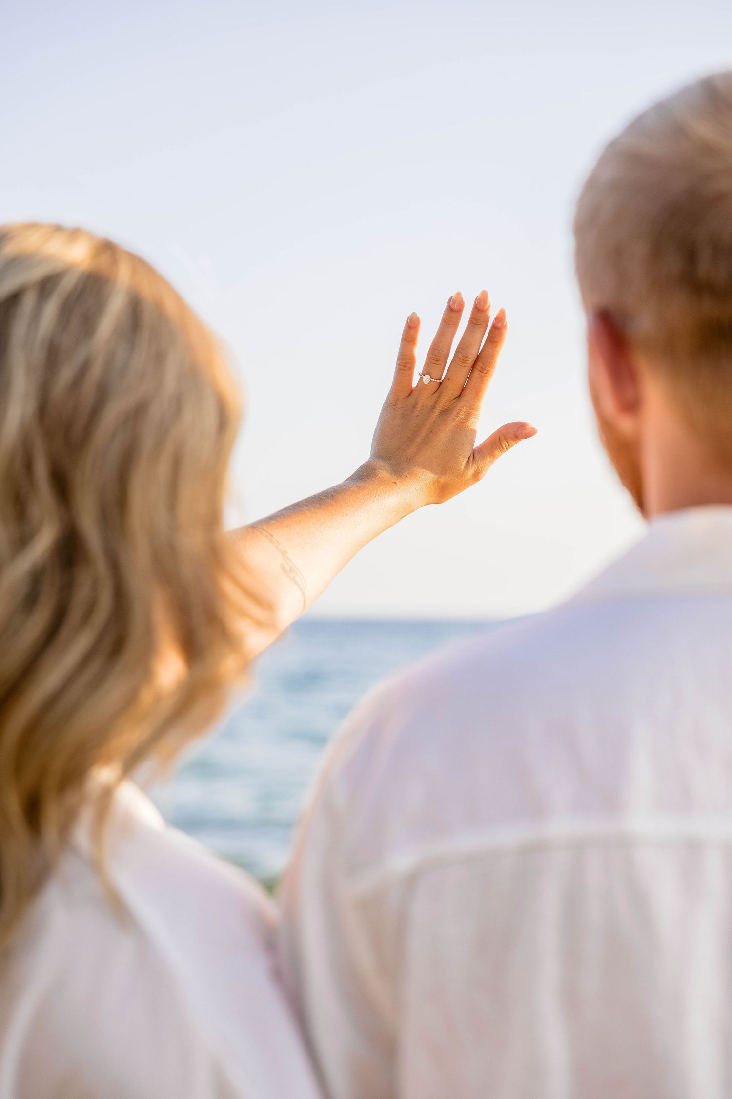 Couple in white linen shirts at beach, woman showing engagement ring, ocean backdrop.