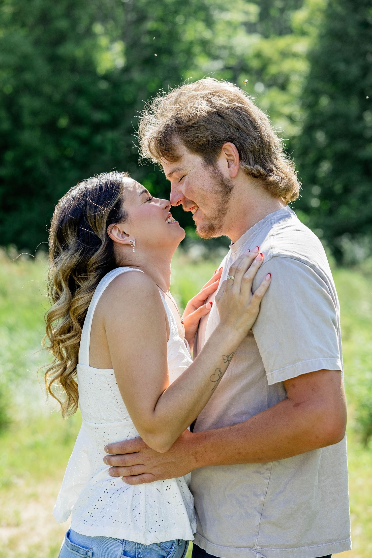 A smiling couple embraces outdoors, nearly kissing, surrounded by lush green trees on a sunny day.