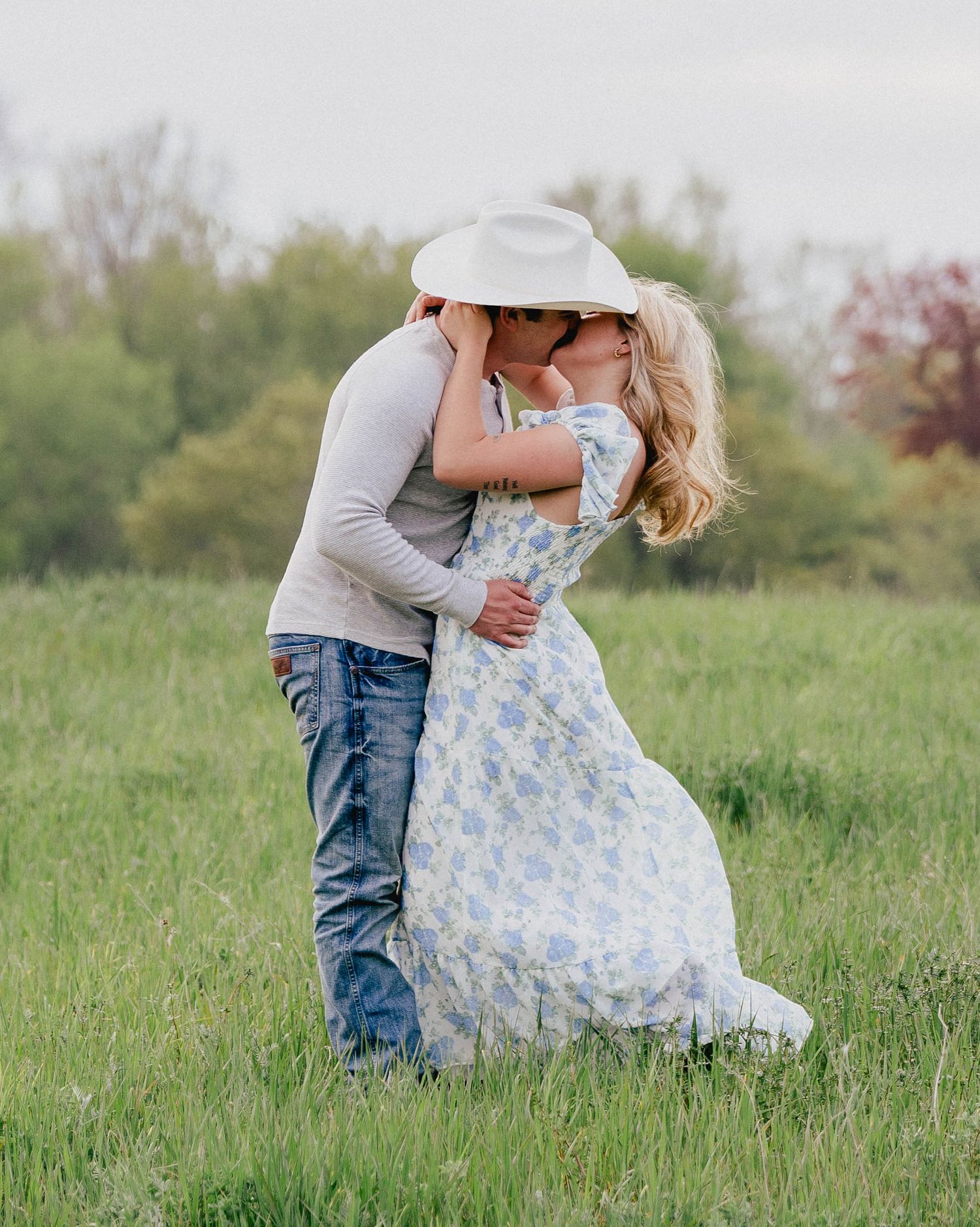 A couple shares a romantic kiss in a lush green field, the man wearing a white cowboy hat.