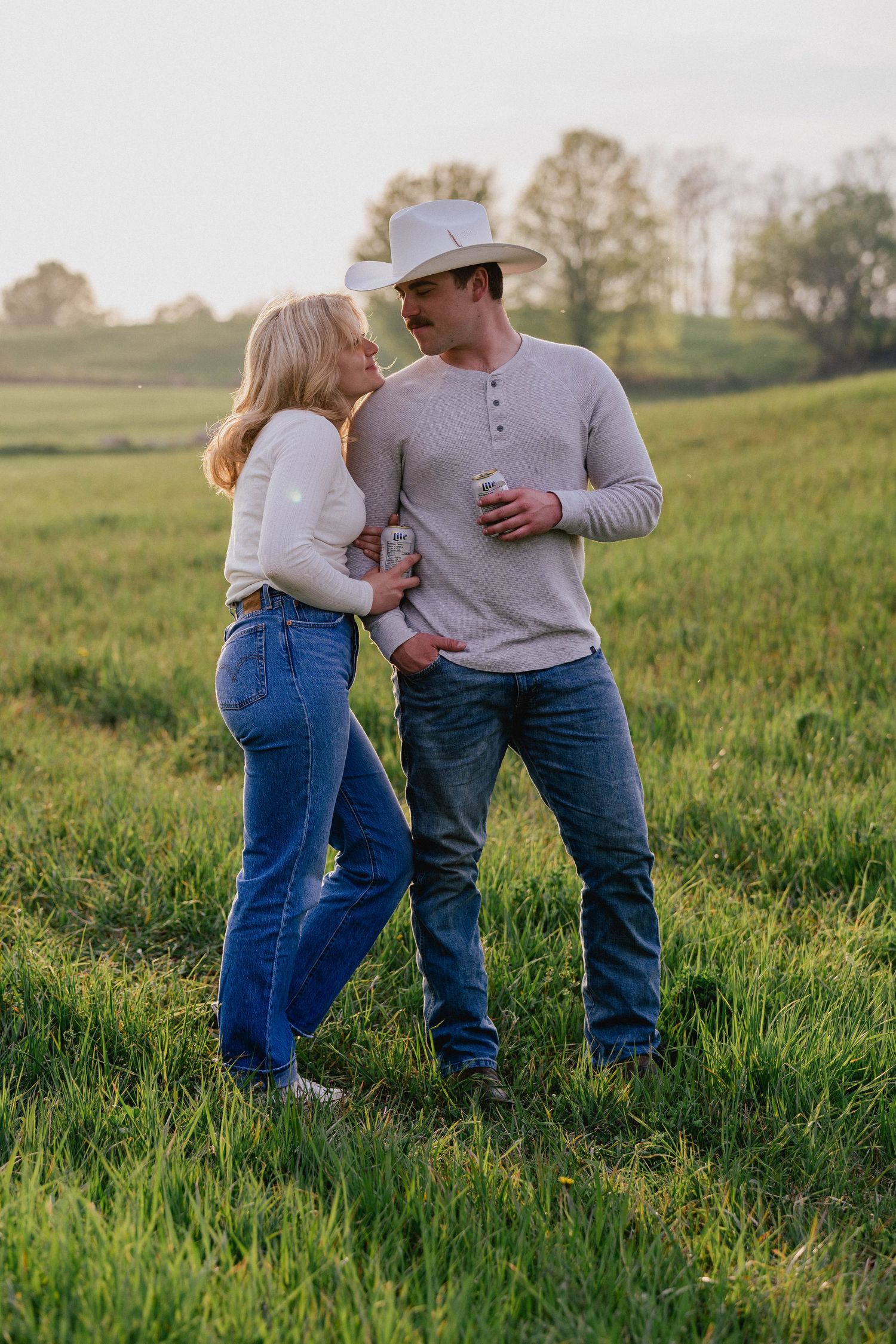 Couple in jeans and cowboy hat sharing a romantic moment in a lush green field at sunset.