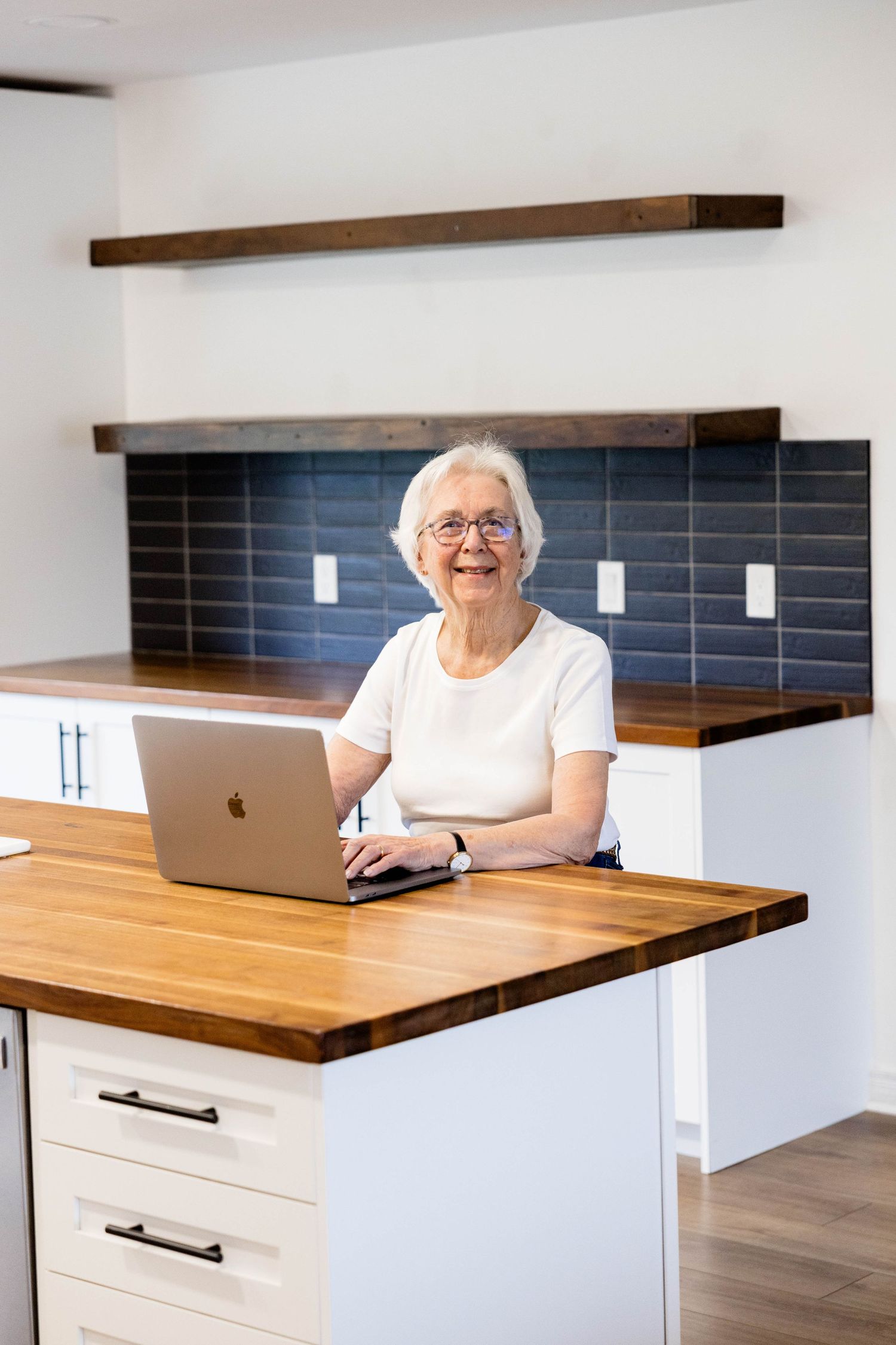 Senior woman working on laptop at kitchen island with butcher block counter and blue tile backsplash.