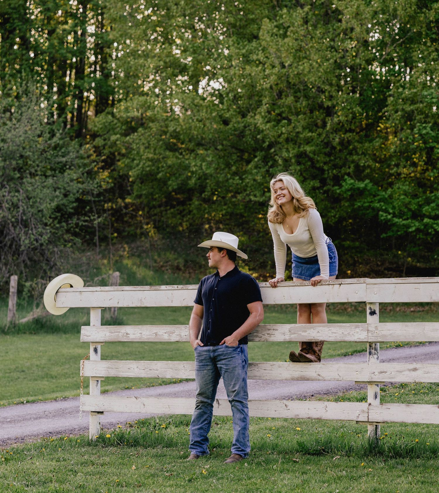 Cowboy in white hat chats with smiling blonde woman perched on a white wooden fence.