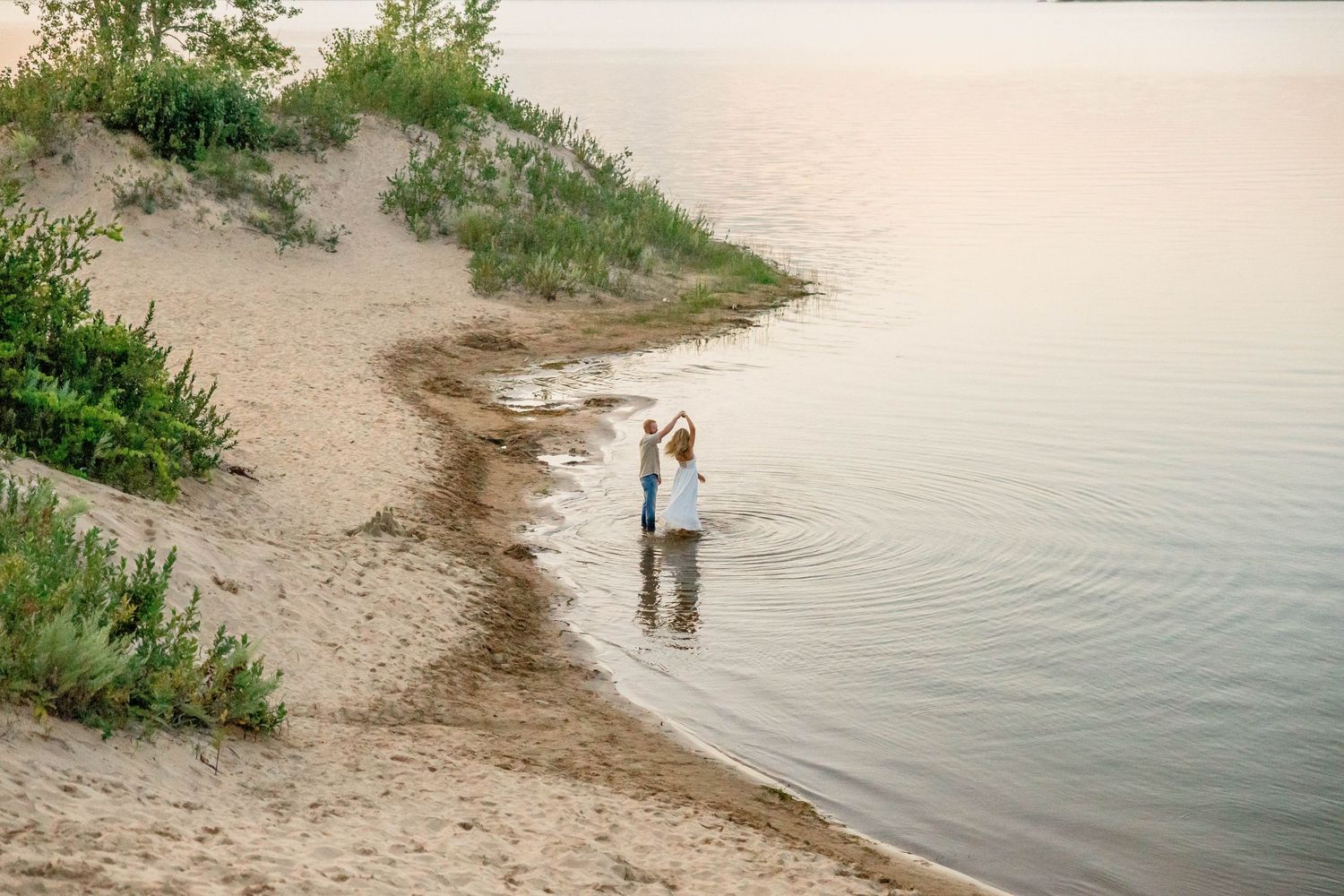Couple standing in shallow water at a sandy beach shoreline during golden hour sunset.
