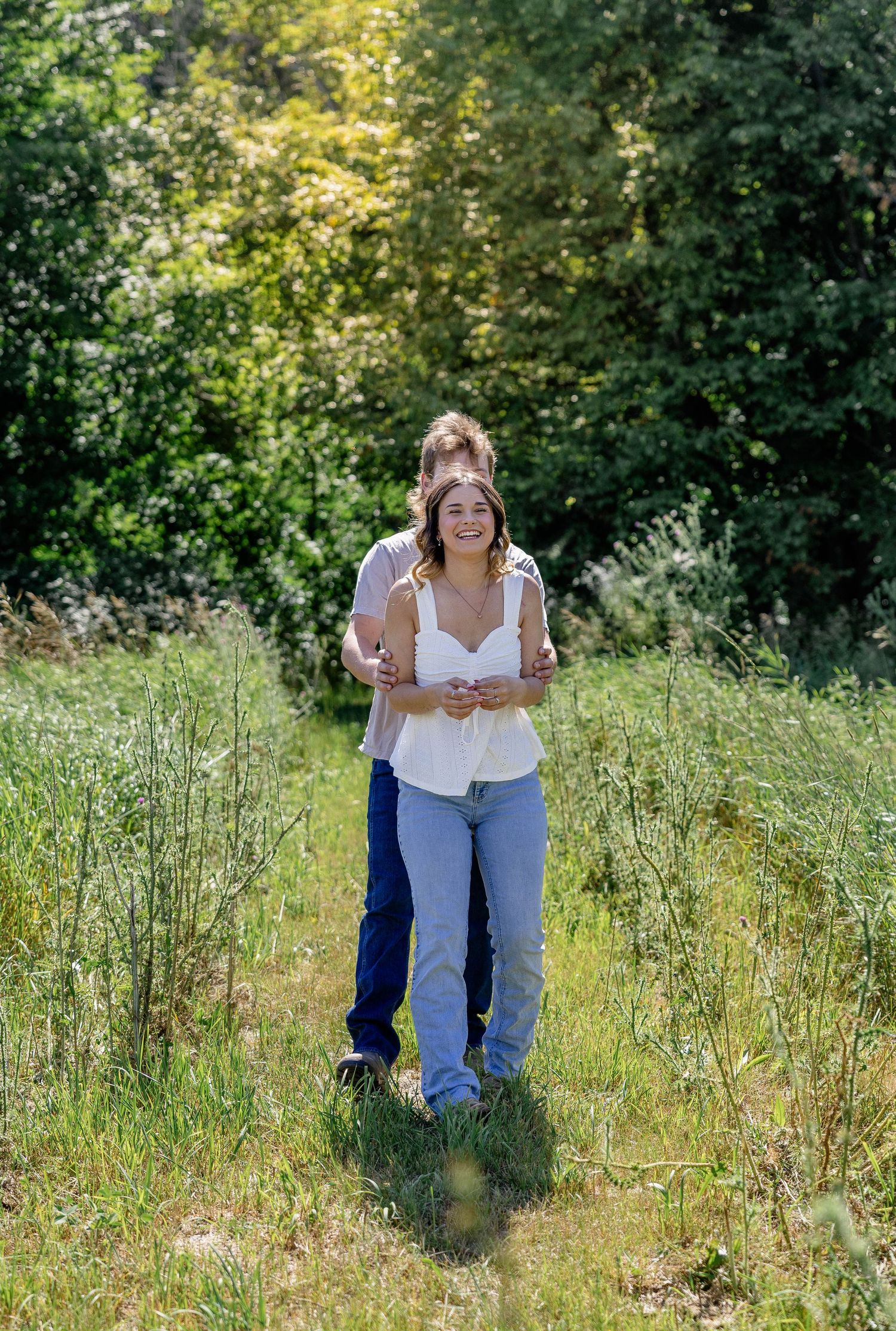 Couple embracing and walking through a lush green meadow on a sunny day, woman in white tank top and jeans smiling.