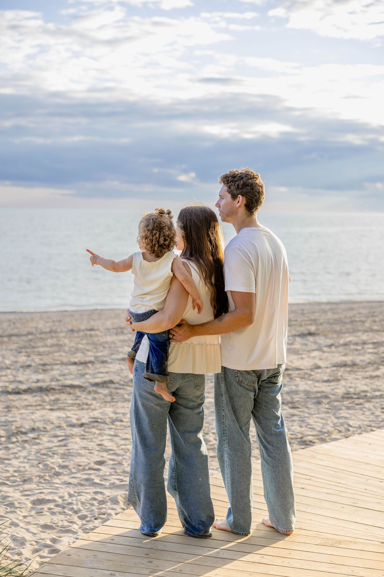 Young family of three standing on a beach boardwalk, child pointing at the ocean horizon at sunset.