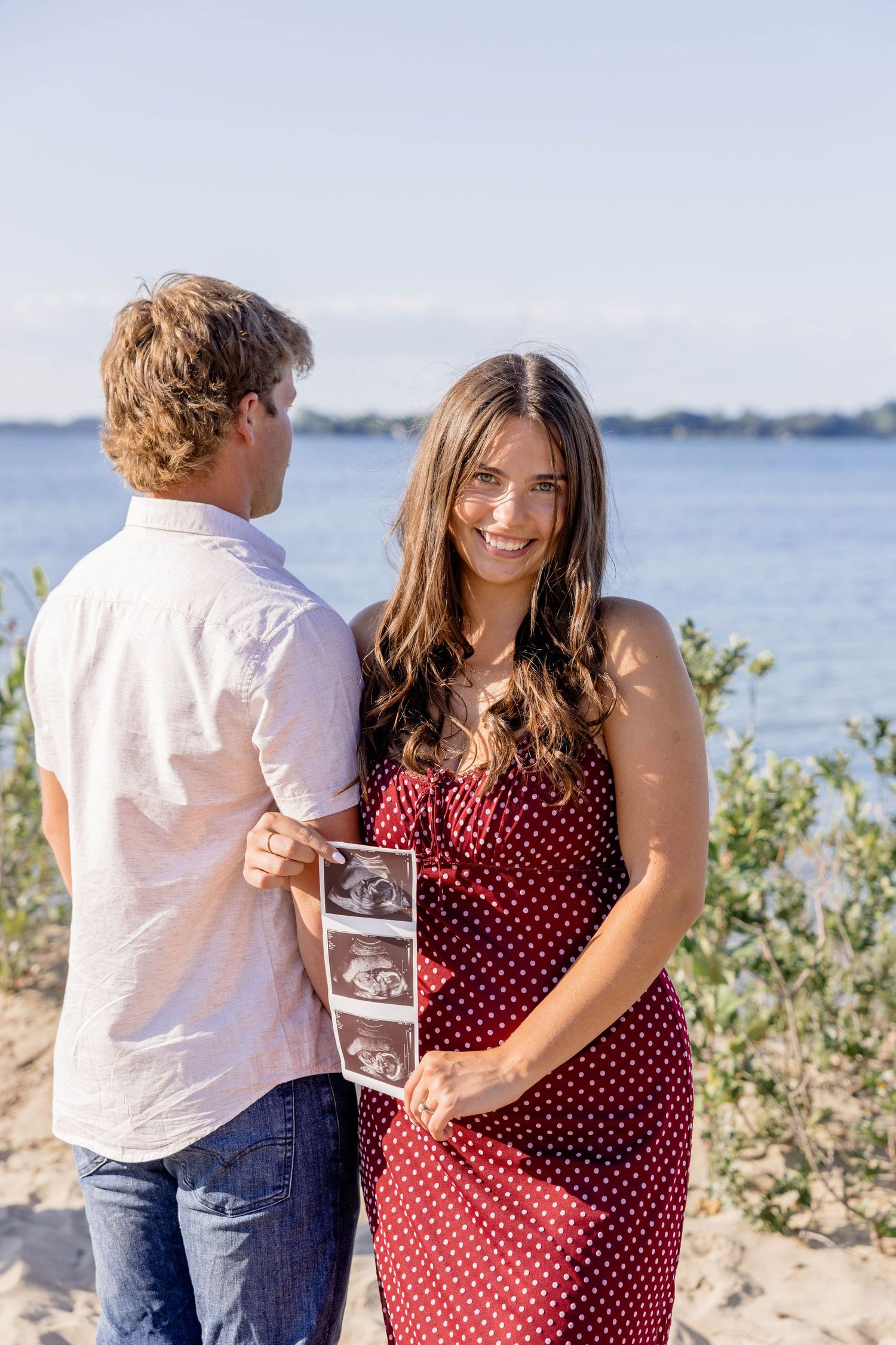Smiling pregnant woman in red polka dot dress holds ultrasound photos with partner on sunny beach.