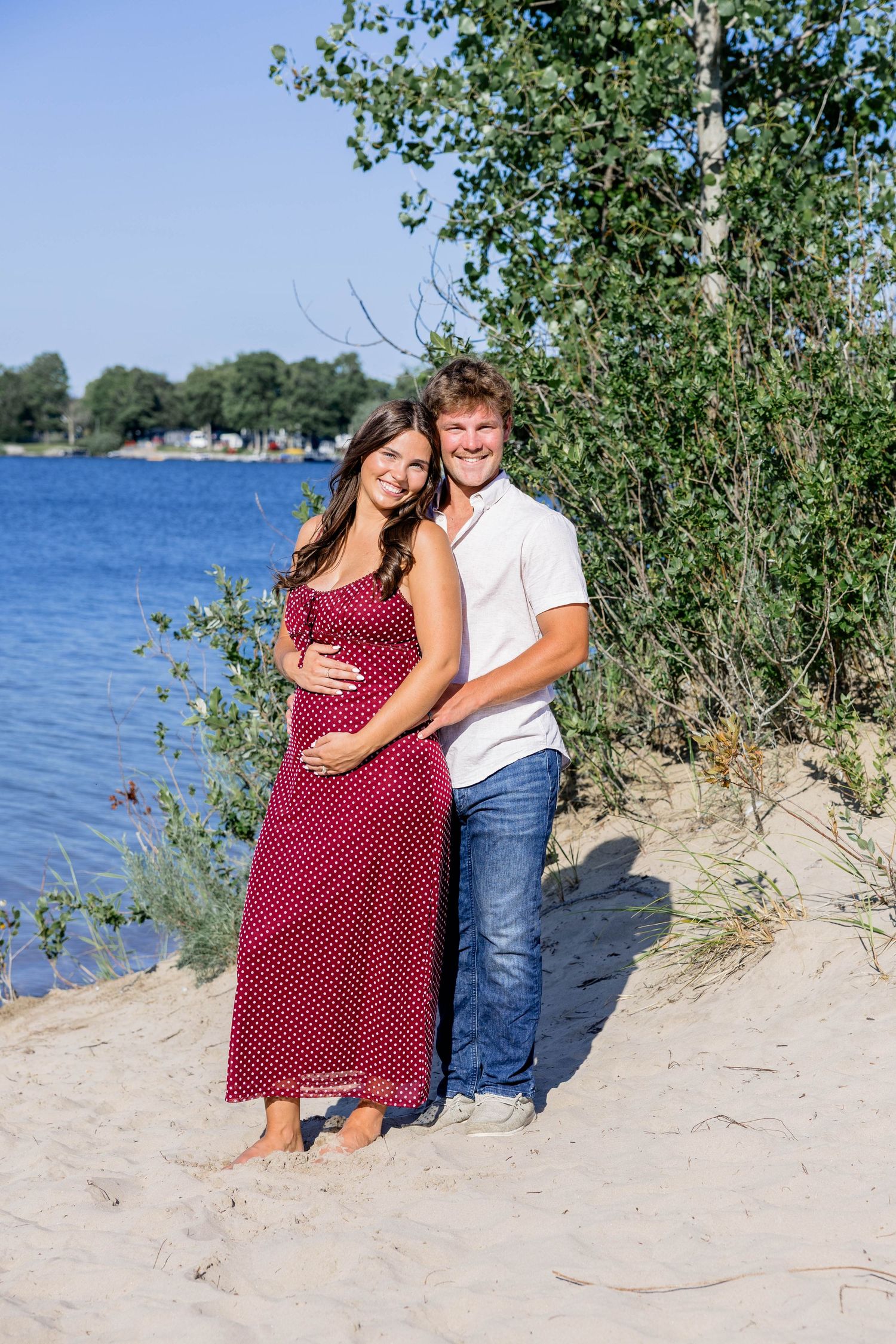 Smiling pregnant couple embracing on sandy beach by blue lake, woman in red polka dot dress, man in white shirt and jeans.