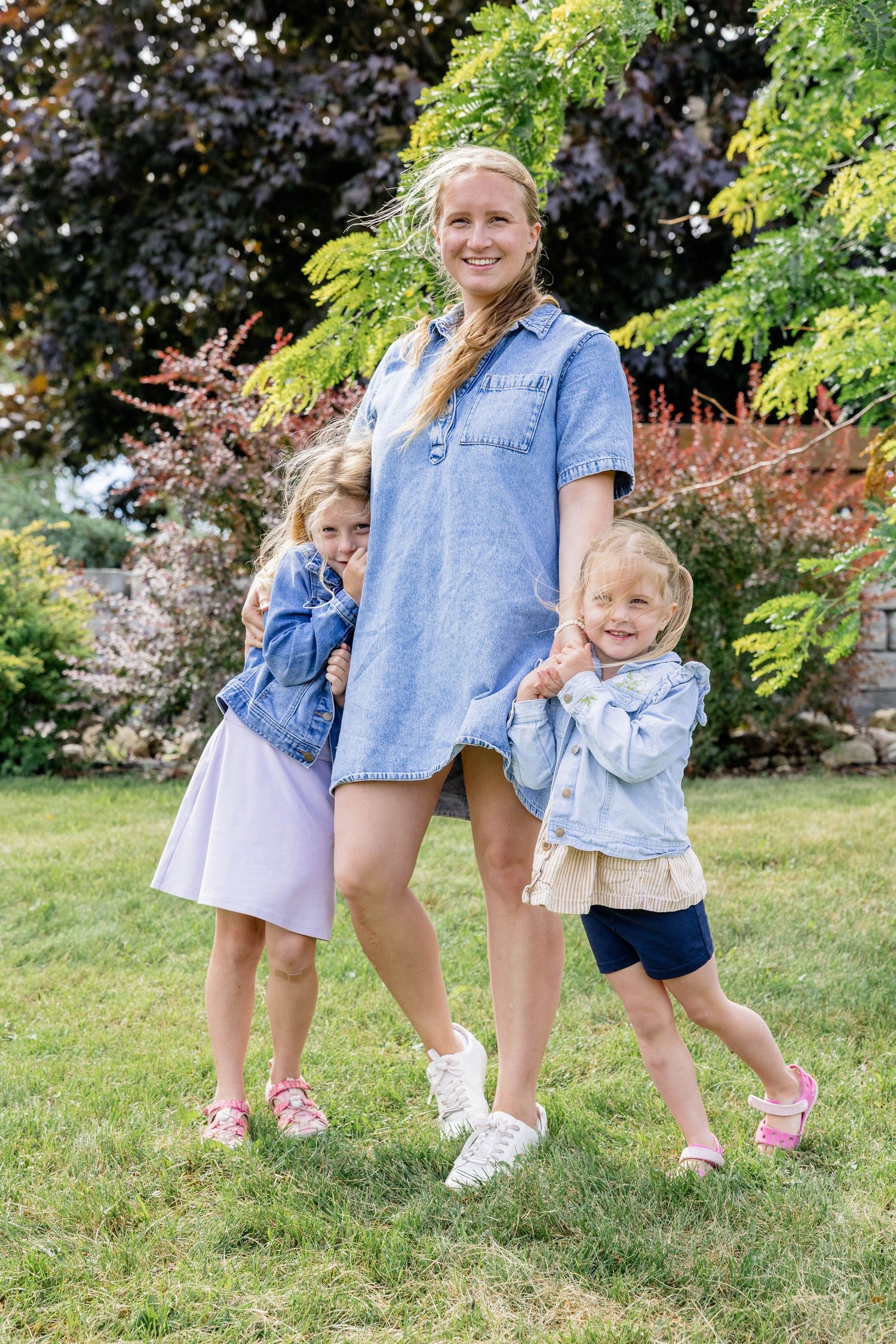 A smiling woman in a denim dress poses outdoors with two young girls in denim jackets on a green lawn.