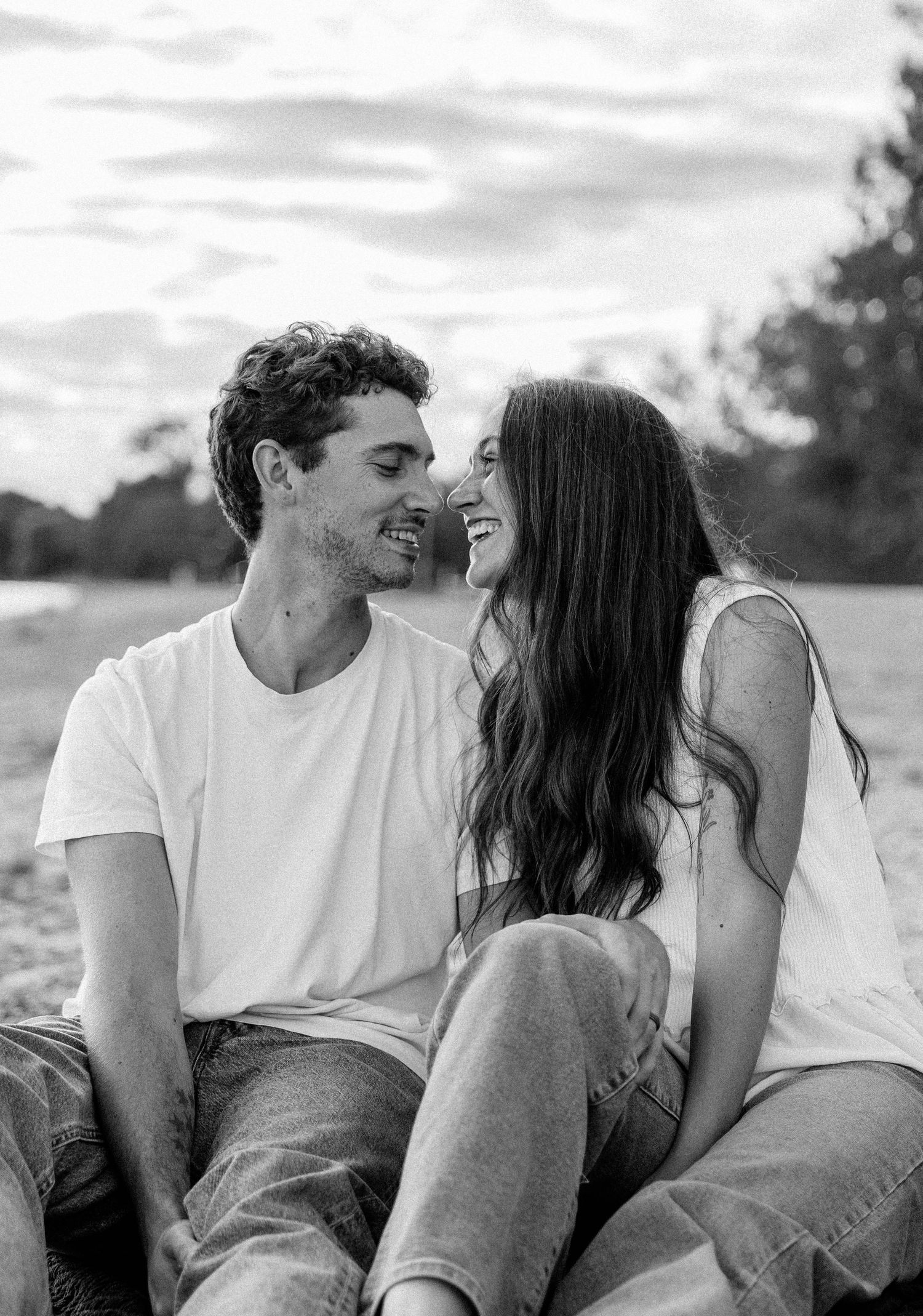 Black and white photo of a smiling couple sitting closely together outdoors, gazing at each other romantically.