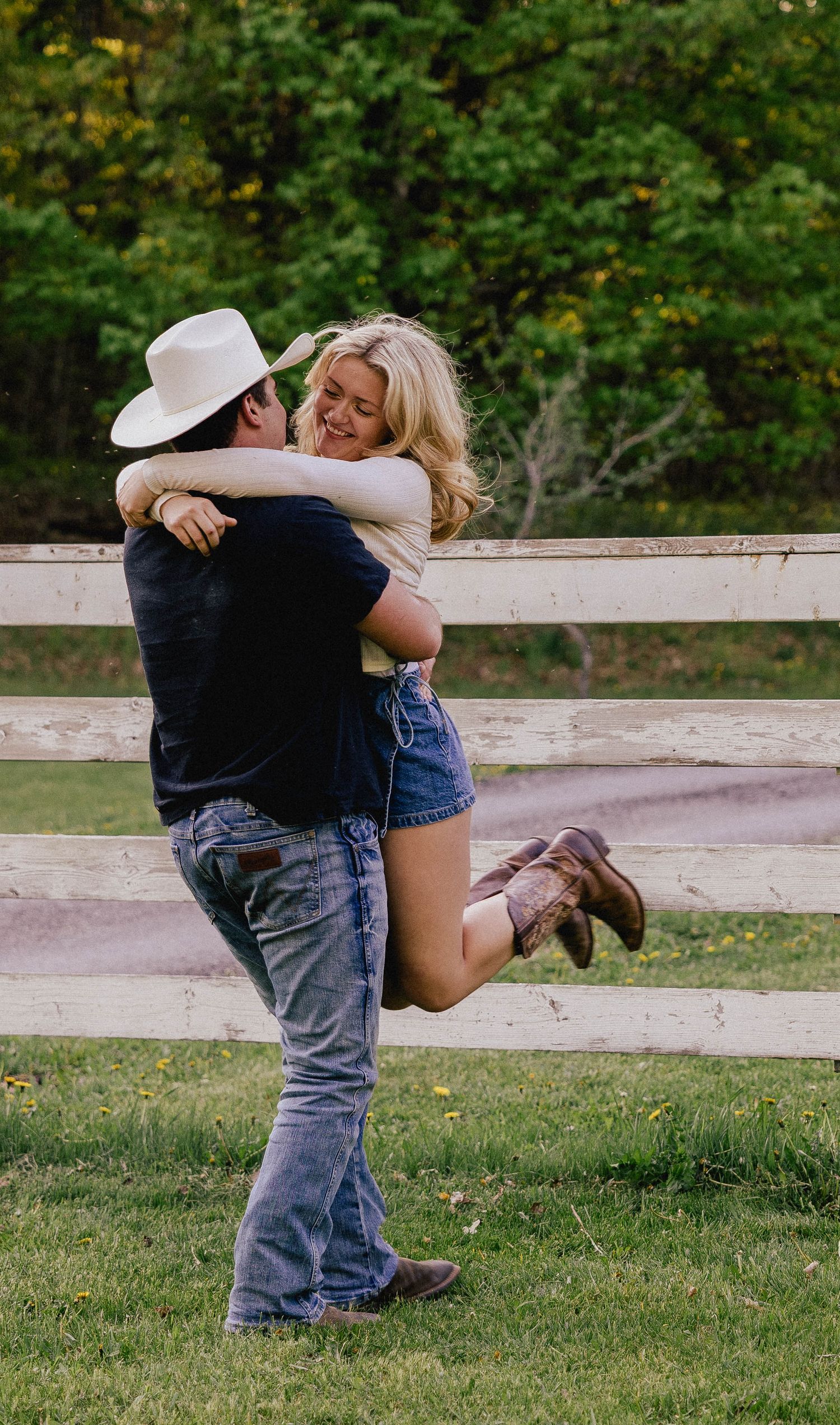 Man in cowboy hat lifting and spinning smiling woman outdoors near a white wooden fence.