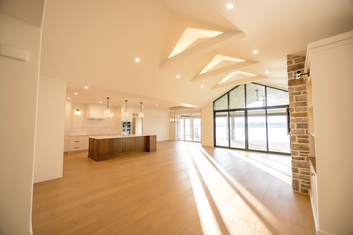 Expansive open-plan living area with vaulted ceiling, kitchen island, stone pillar, and large windows.