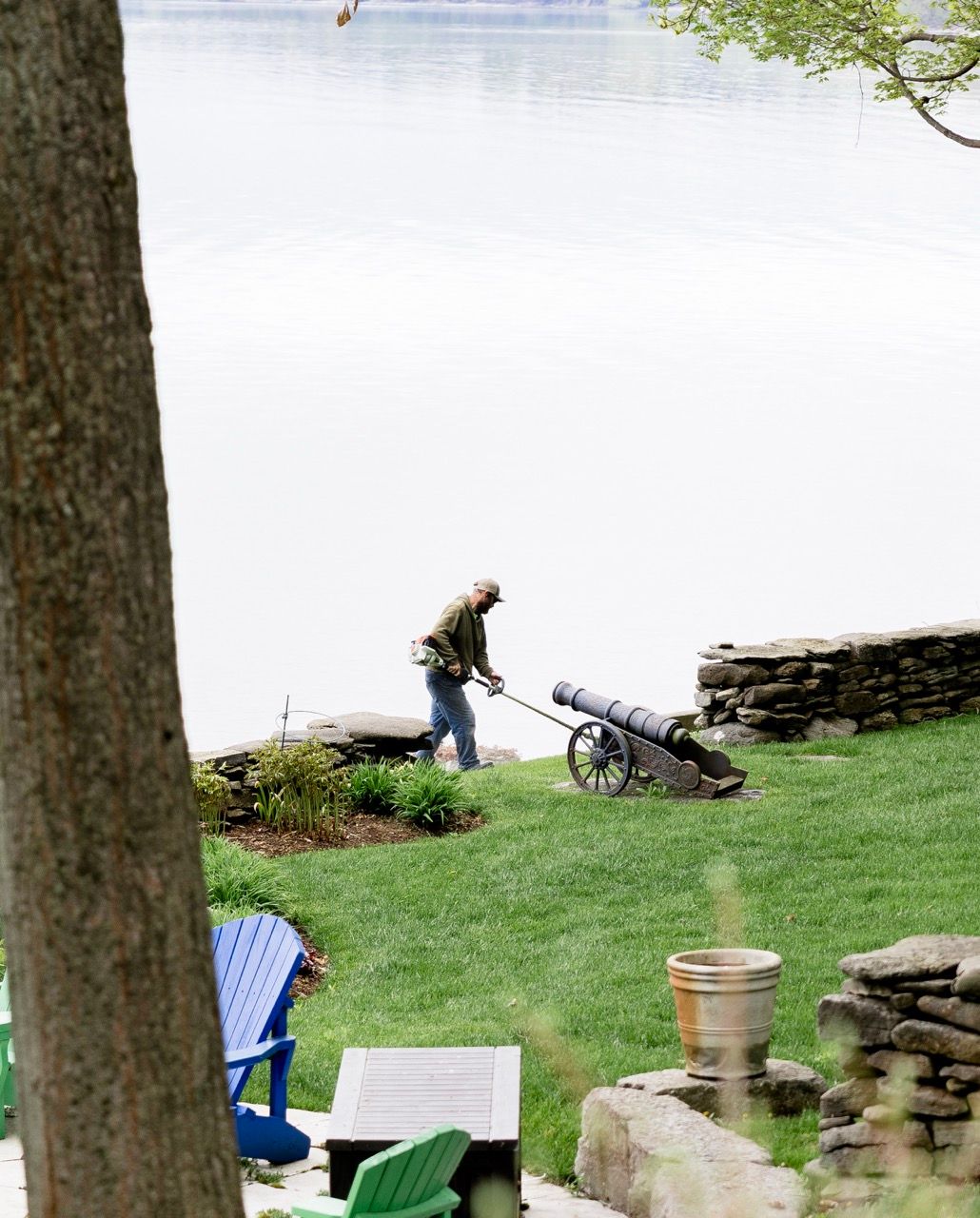 Man using a long-handled tool near a decorative cannon by a lakeside stone wall garden.