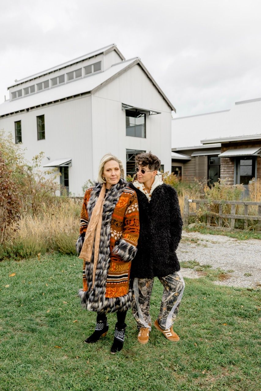 Two stylish individuals pose together on a lawn with a modern white barn-style building behind them.