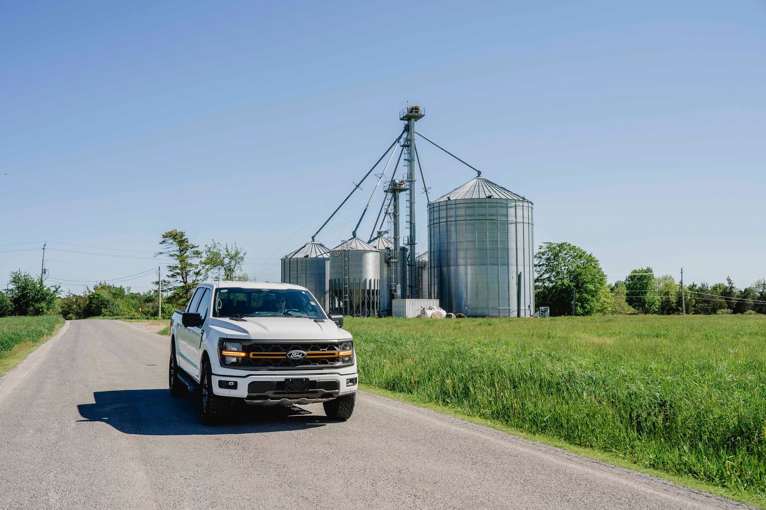 White Chevrolet pickup truck parked on rural road beside grain silos and green fields.