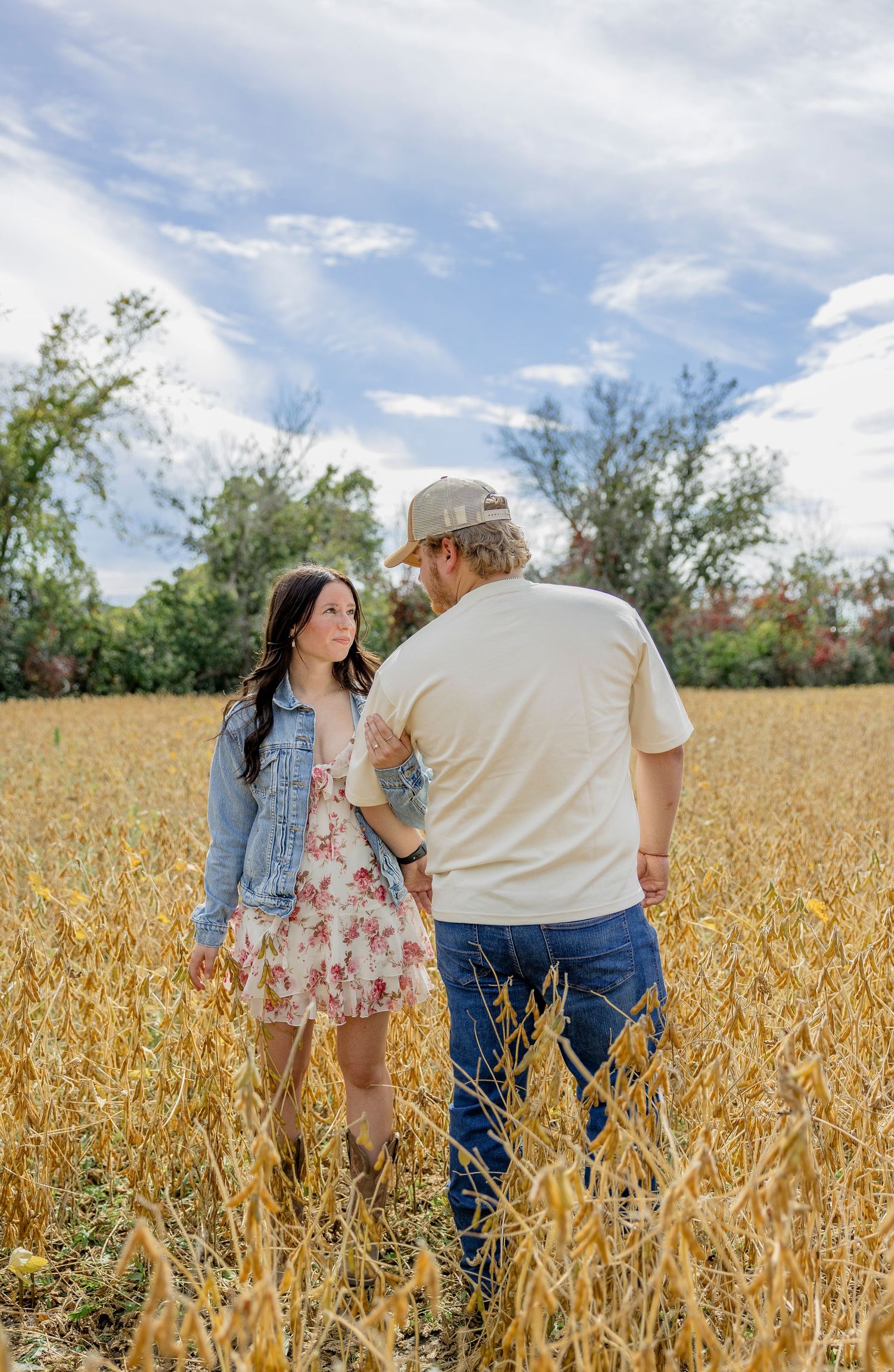 Couple walking through golden harvest field, woman in floral dress and denim vest, man in cap and cream shirt, under cloudy sky.