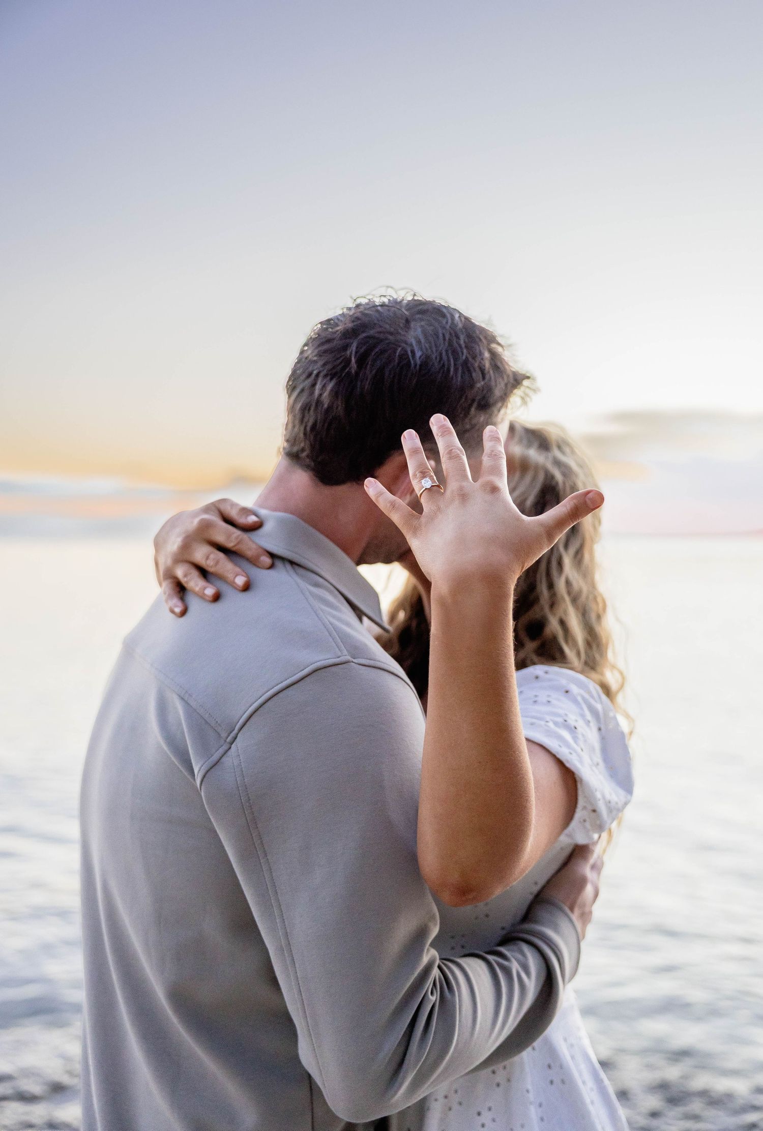 Couple kissing at sunset by the ocean, woman showing off engagement ring on her hand.