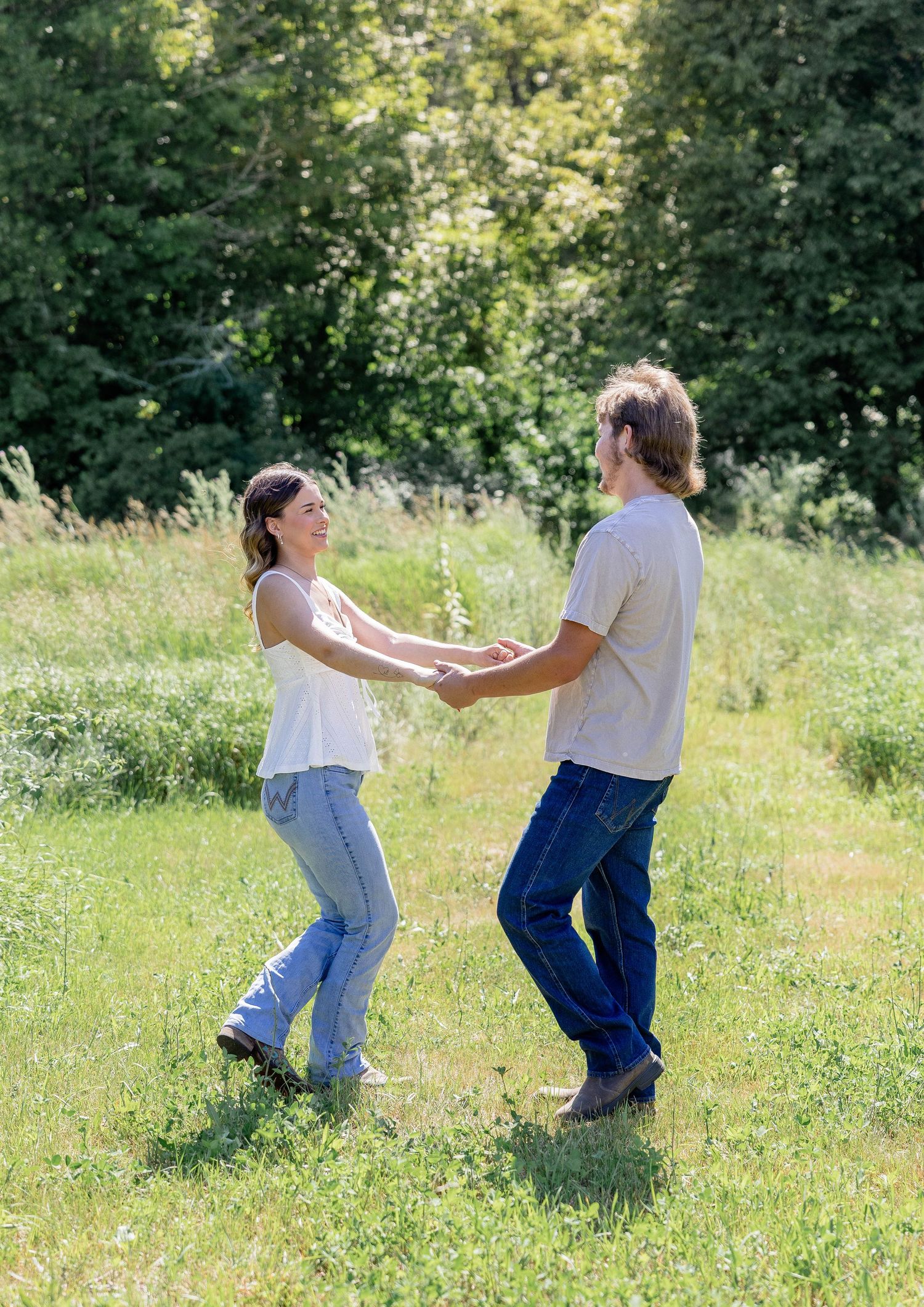 A joyful couple holds hands and dances together in a sunny green meadow surrounded by lush trees.