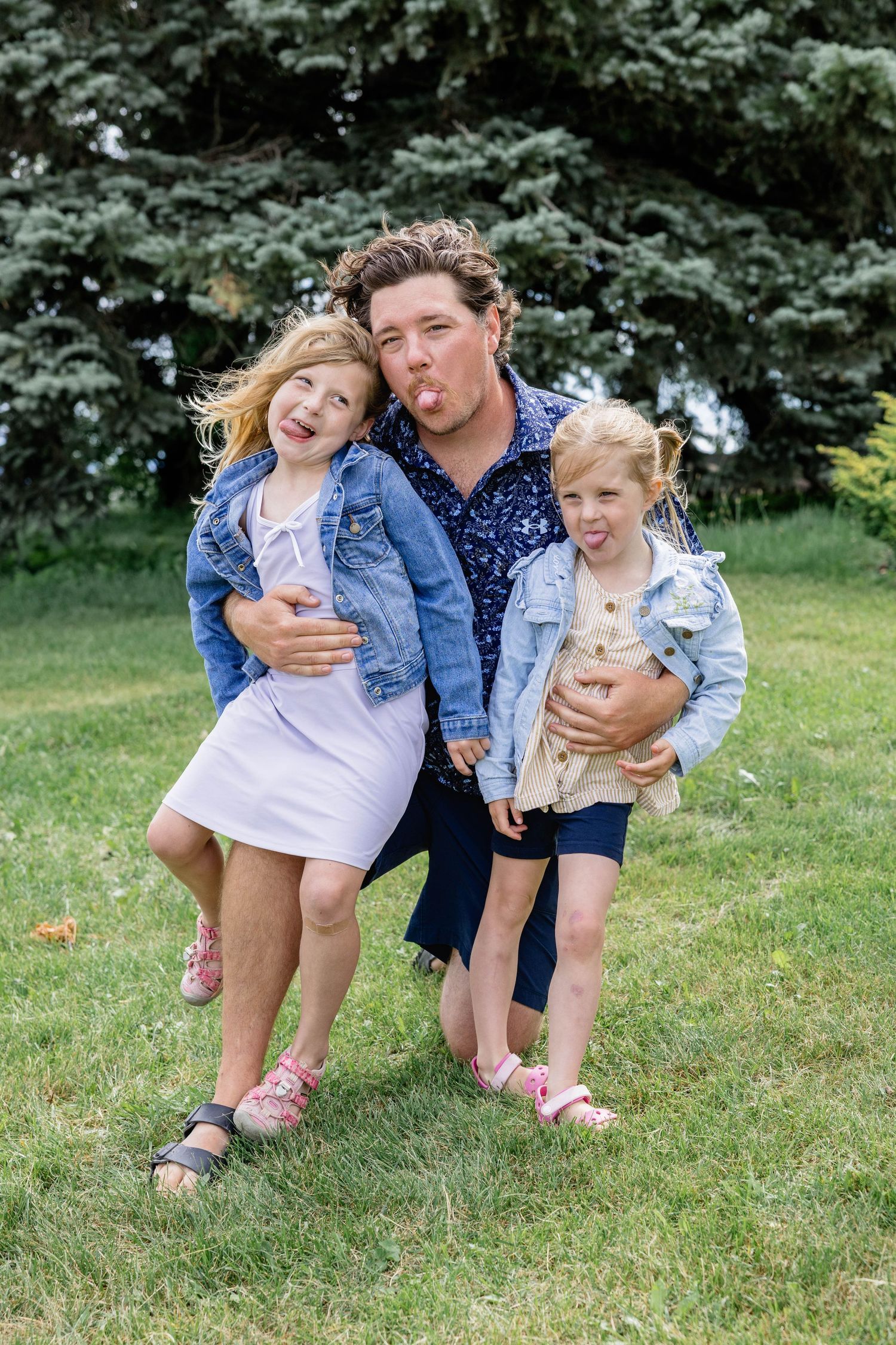 Parent hugging two young children in denim jackets, making silly faces outdoors on green grass with evergreen trees.