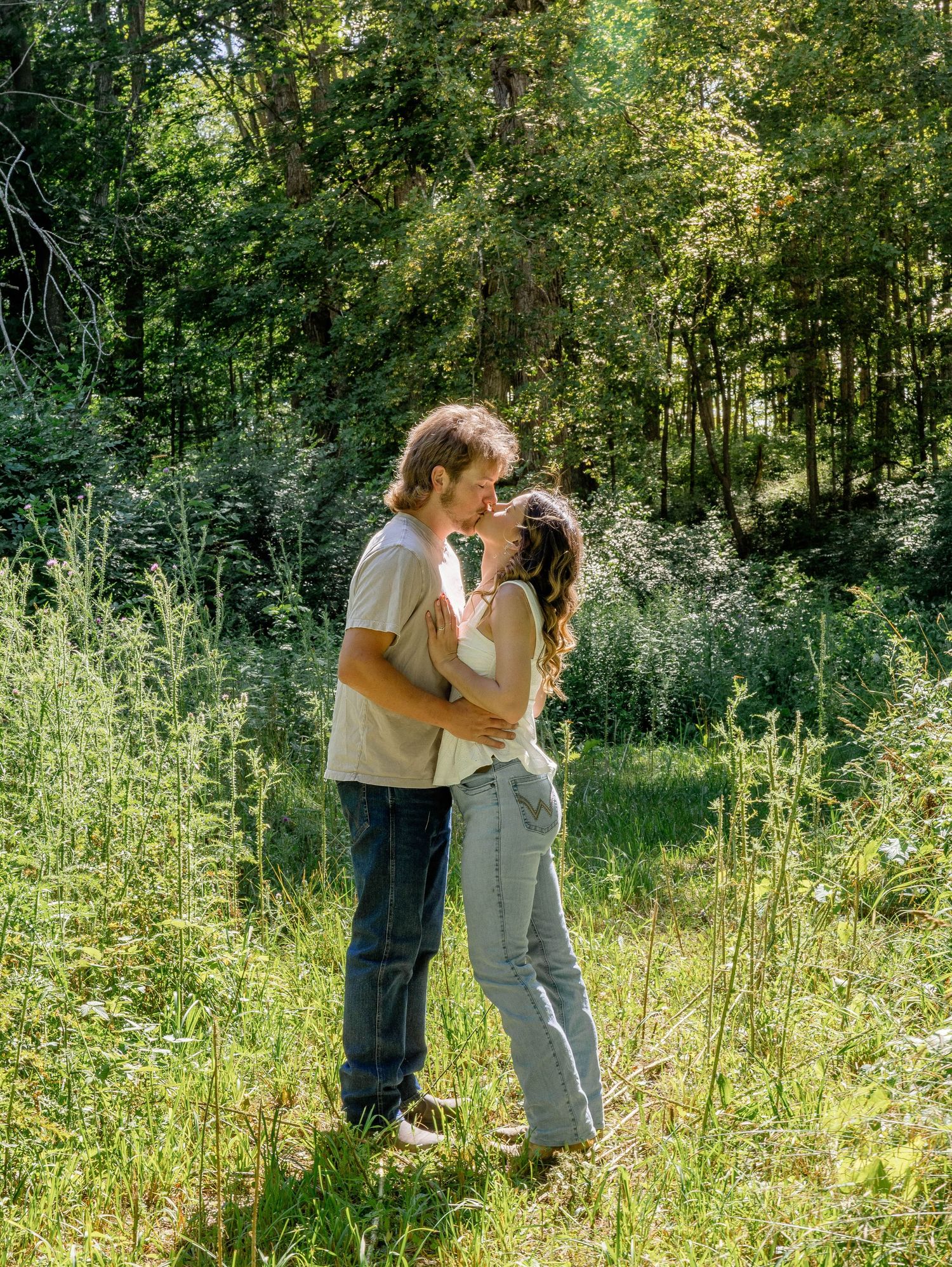 A couple shares a romantic kiss in a lush green meadow surrounded by tall grass and trees on a sunny day.