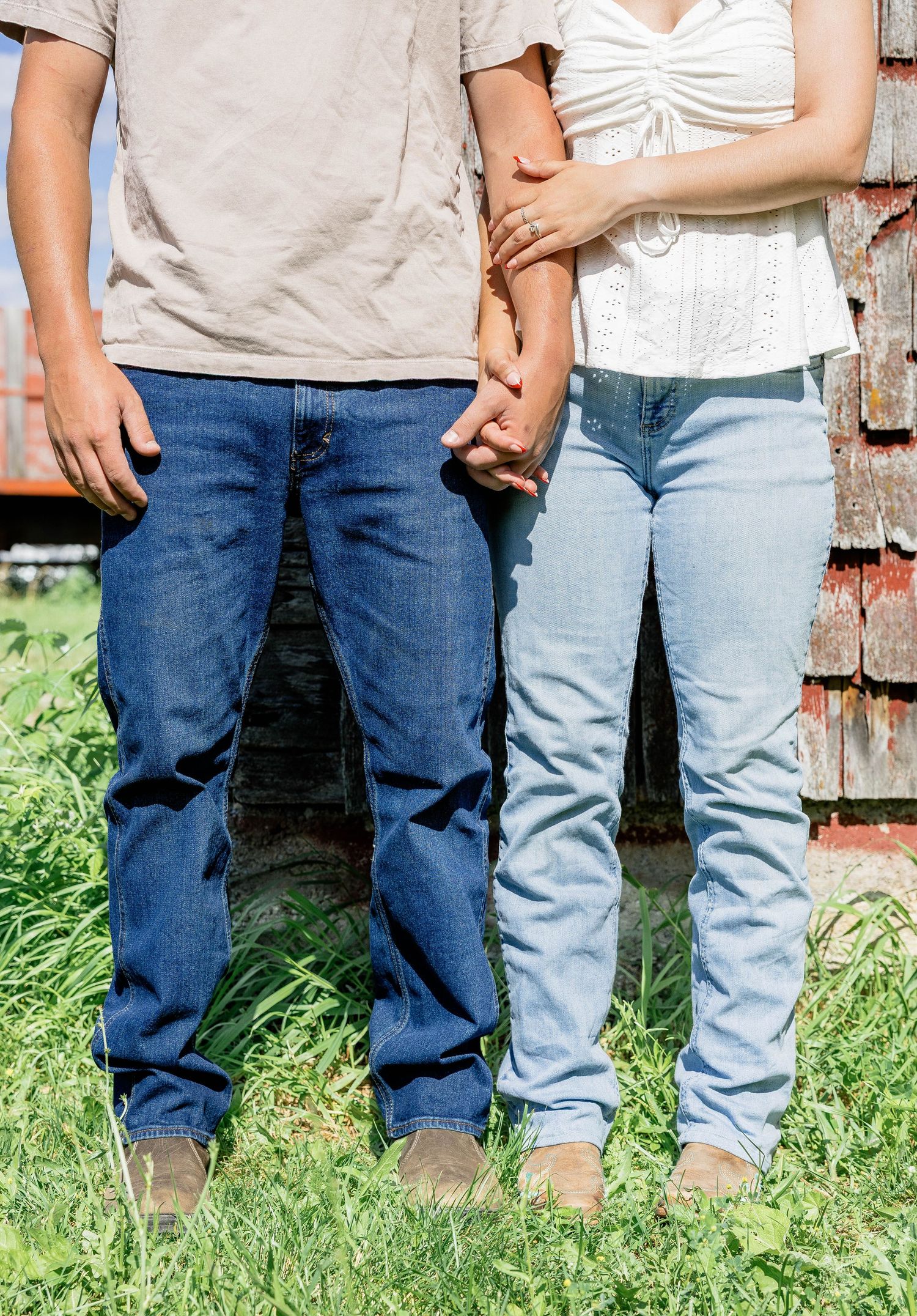 Couple holding hands in jeans standing on green grass near a rustic red barn wall outdoors.