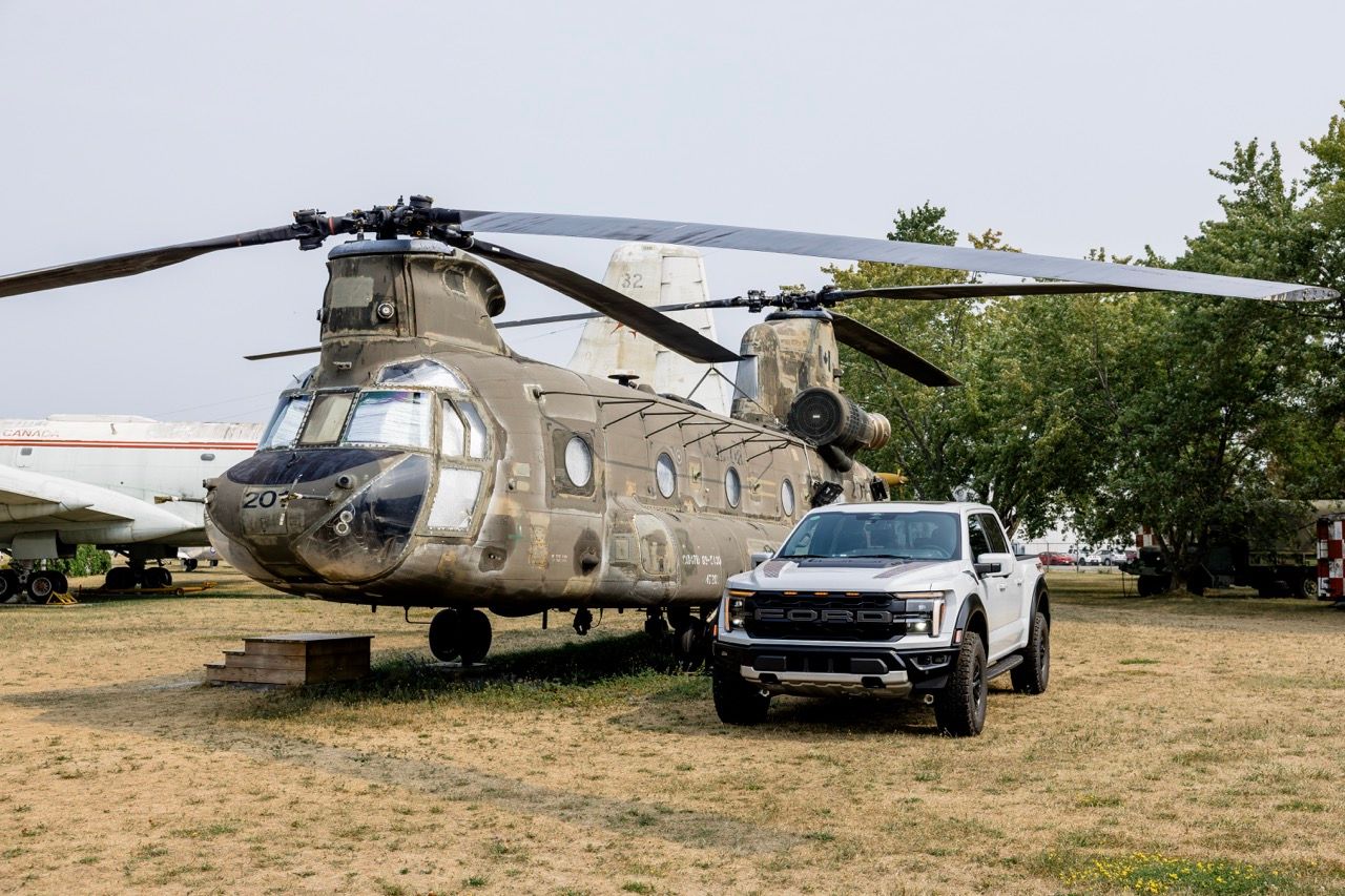 A military CH-47 Chinook helicopter parked beside a white Ford Raptor truck at an outdoor aviation museum.