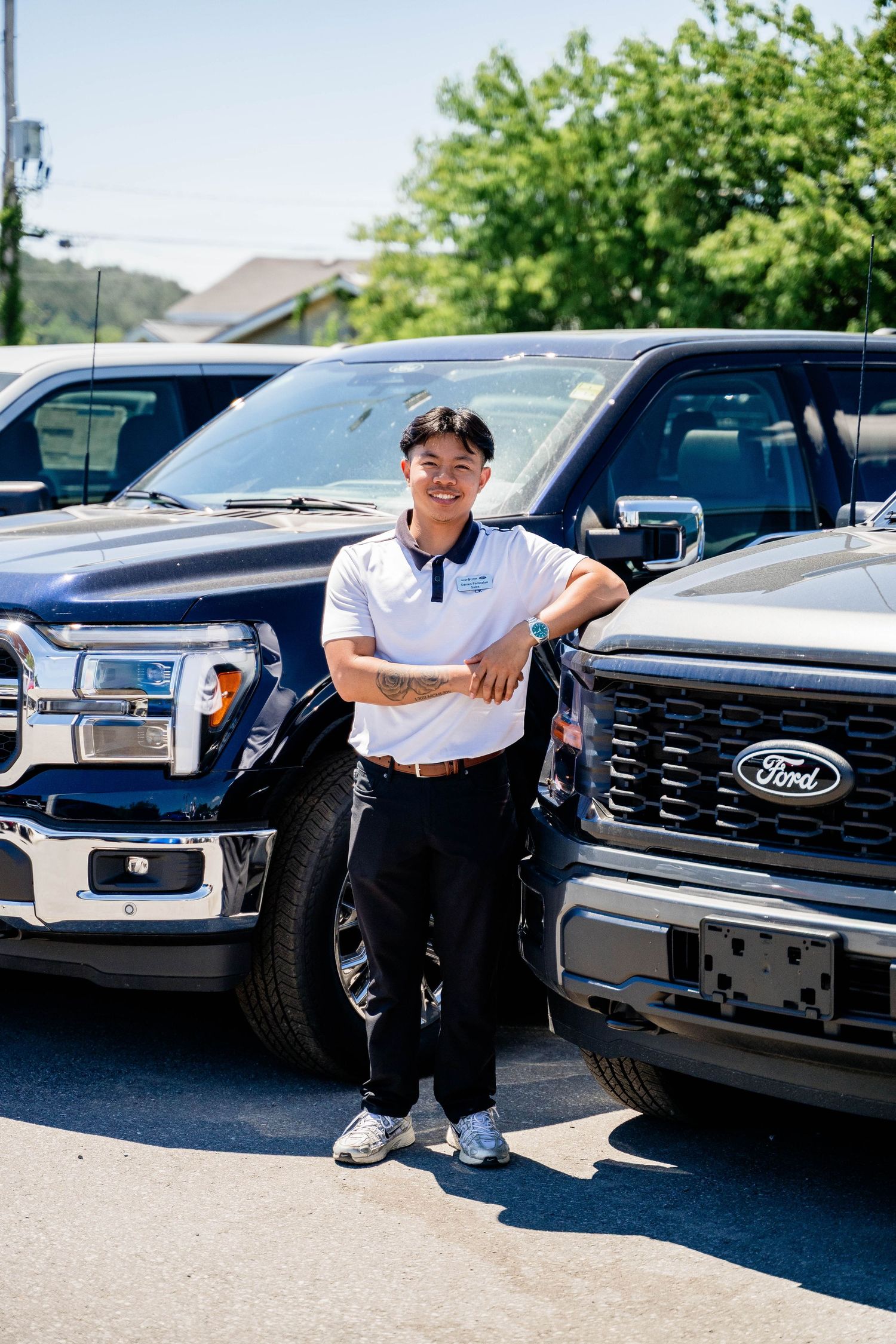 Young man in white polo shirt leaning against Ford F-150 trucks at sunny dealership.