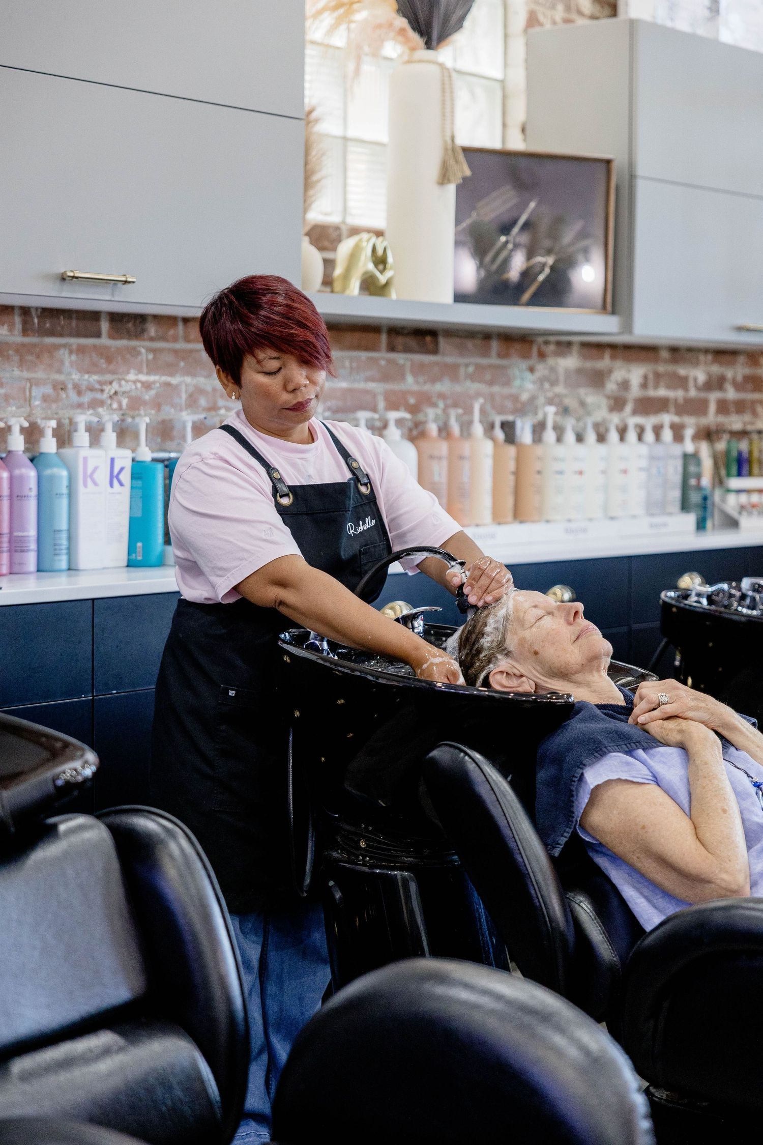 Red-haired stylist washing an elderly client's hair at a salon shampoo bowl.