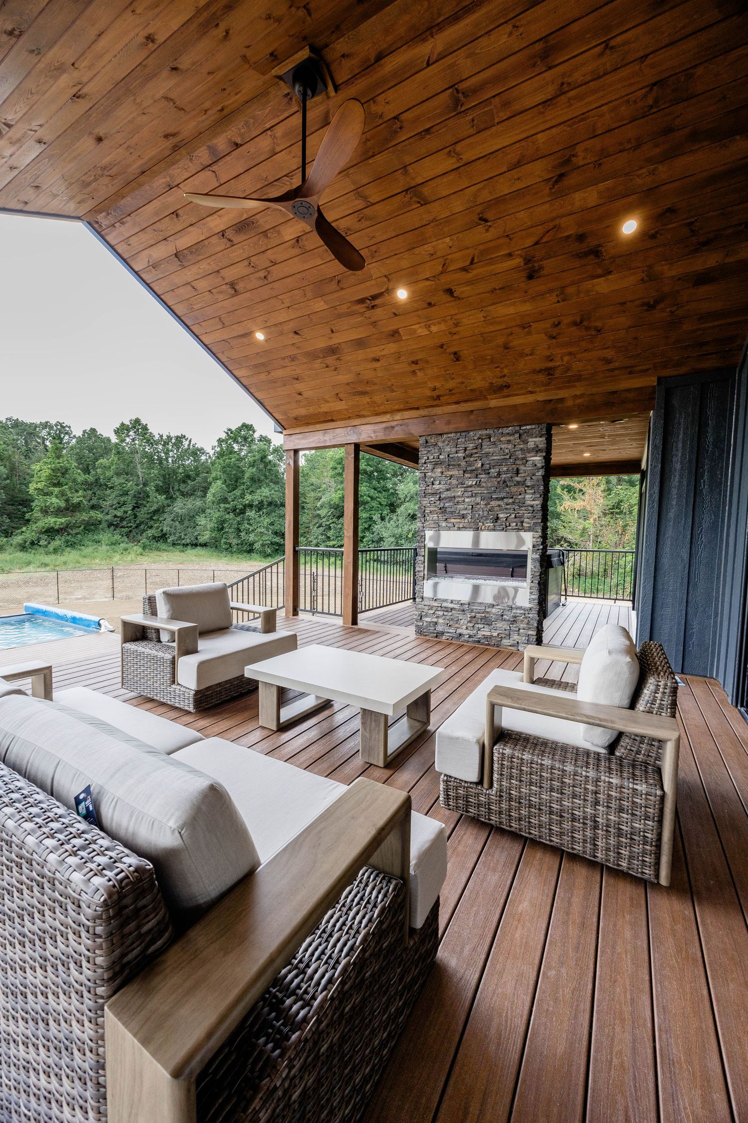 Covered outdoor deck with wicker furniture, stone fireplace, wood ceiling, ceiling fan, and pool view surrounded by lush trees.