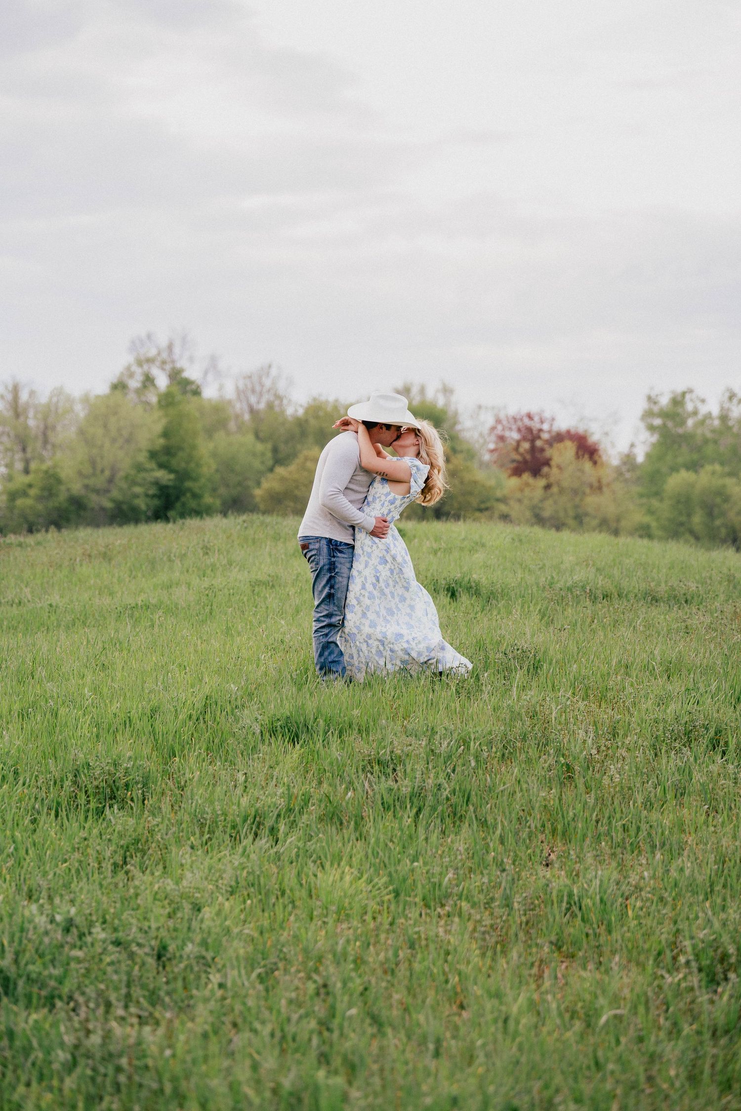 A couple embraces and kisses in a wide open green meadow under a cloudy sky in the countryside.