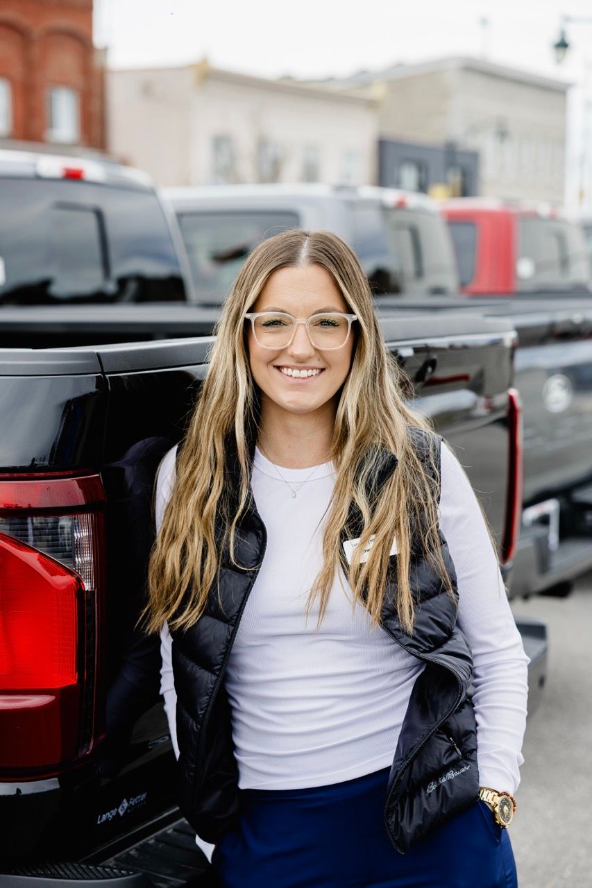 Smiling blonde woman wearing glasses and a black puffer vest posing beside a red truck at a dealership.