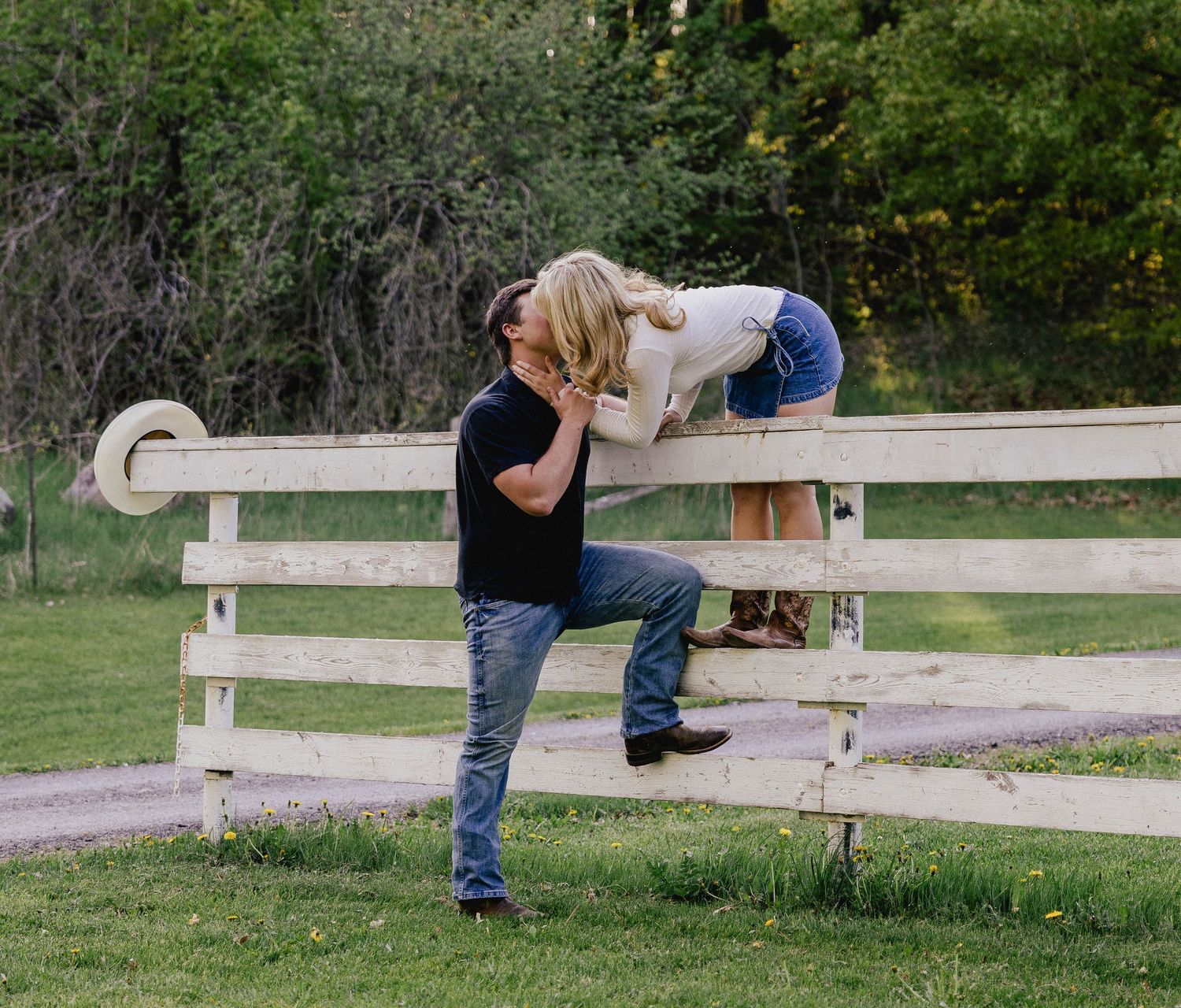 Man in black shirt kisses woman leaning over a white wooden fence in a green field.