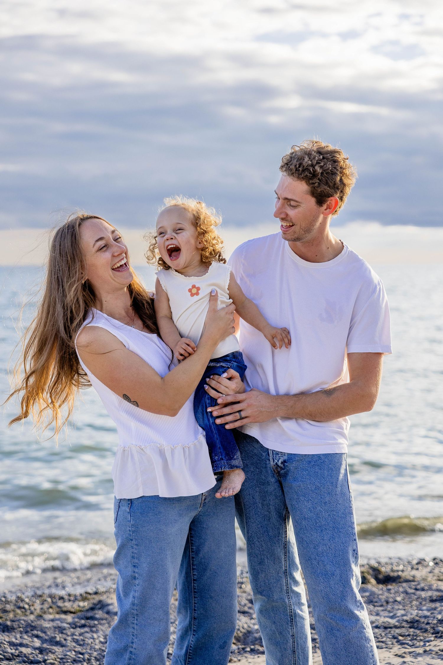 Happy family of three laughing on a rocky beach, parents in white tops and jeans holding their toddler.