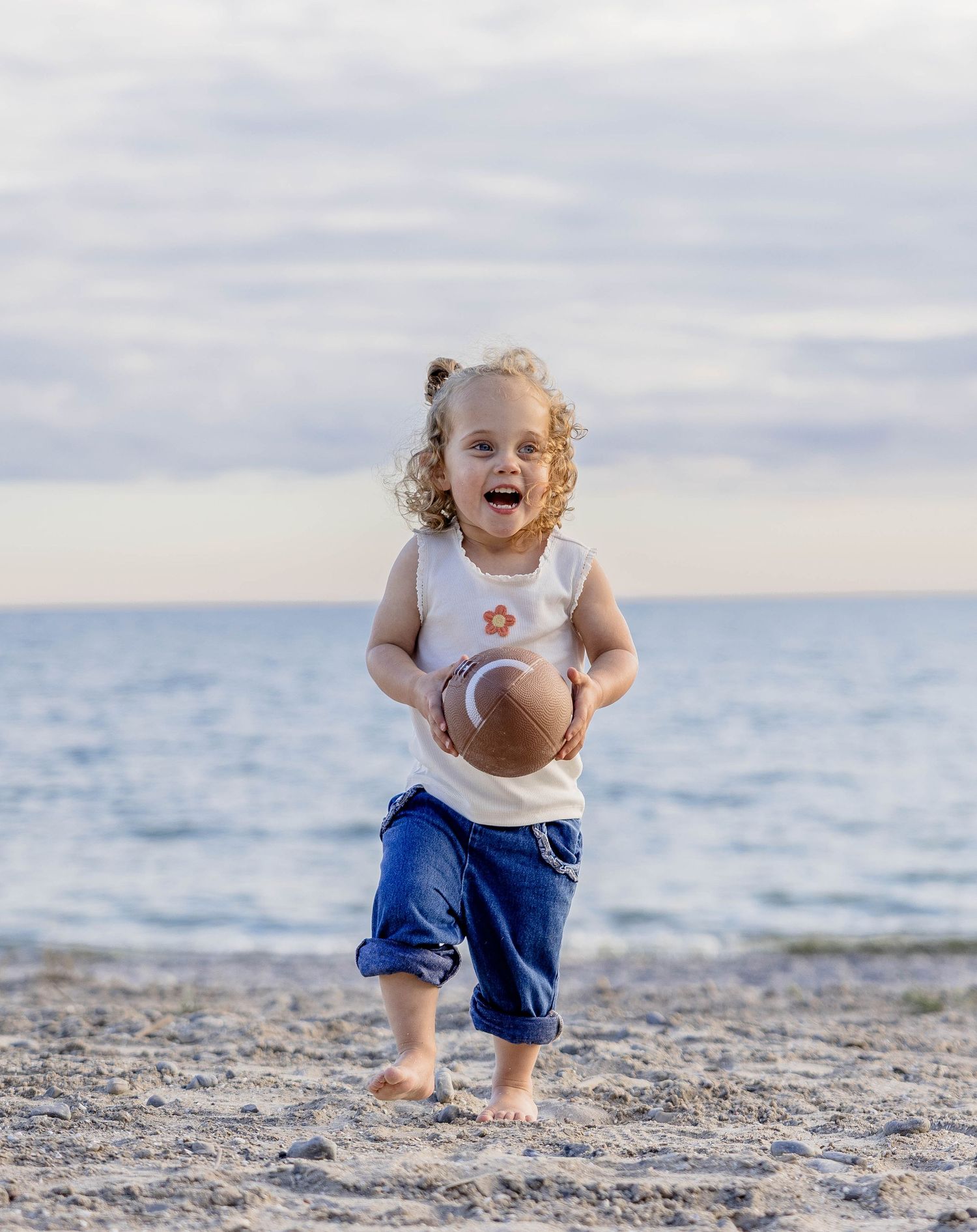 A joyful toddler runs barefoot on a sandy beach holding a football, wearing a white tank top and blue jeans.