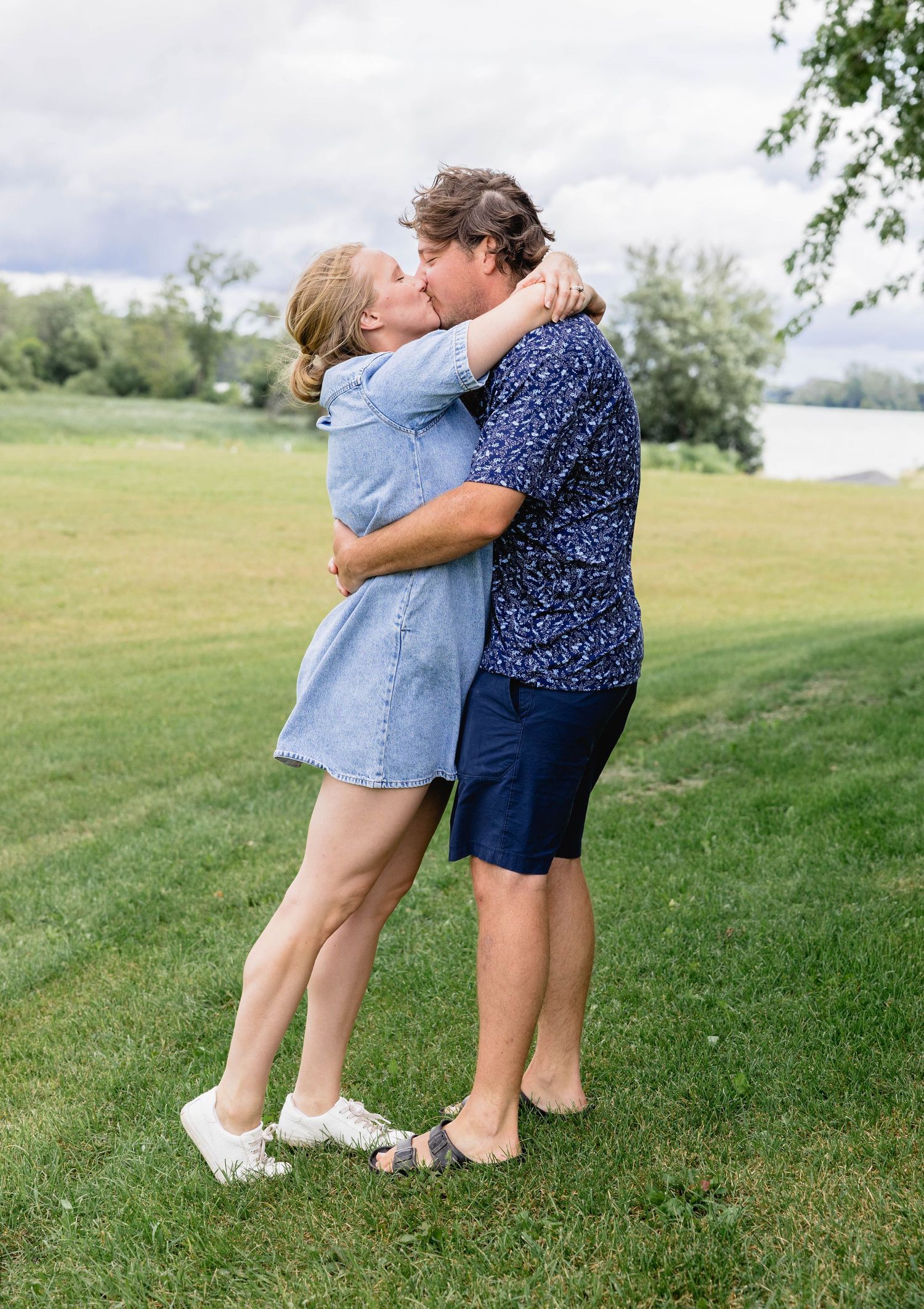 Couple sharing a romantic kiss while embracing on a green lawn near a lake on a cloudy day.