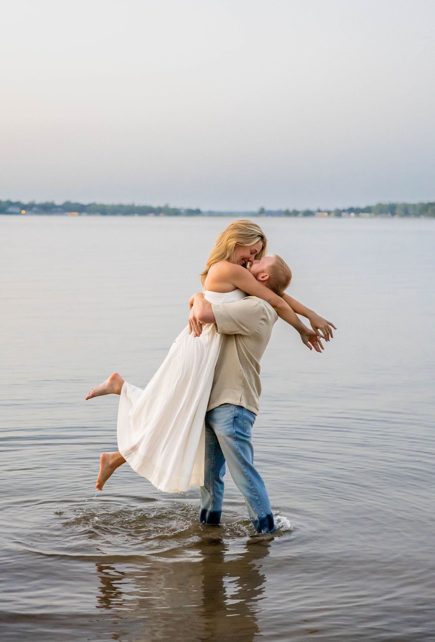 Couple sharing a romantic embrace while wading in shallow lake water at sunset, woman in white dress lifted off ground.