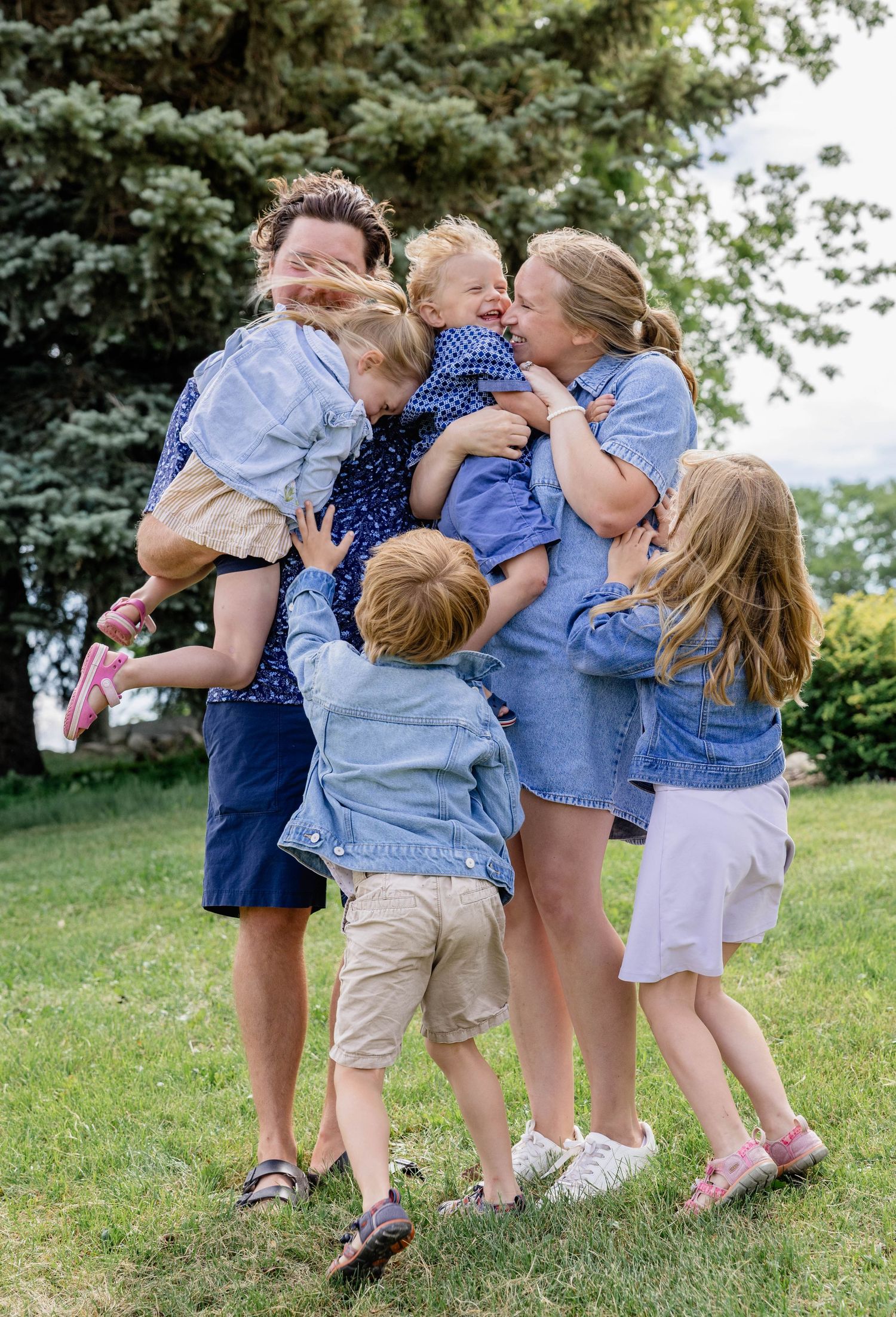 A joyful family of two parents and four young children hugging and laughing together outdoors on green grass.