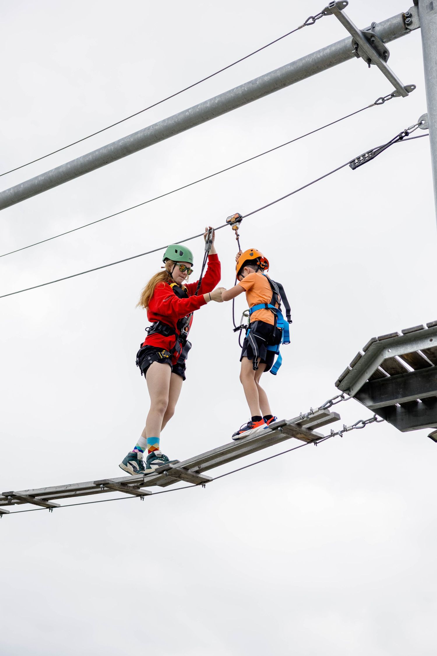 Two people wearing helmets and harnesses walking a high ropes course bridge against a cloudy sky.